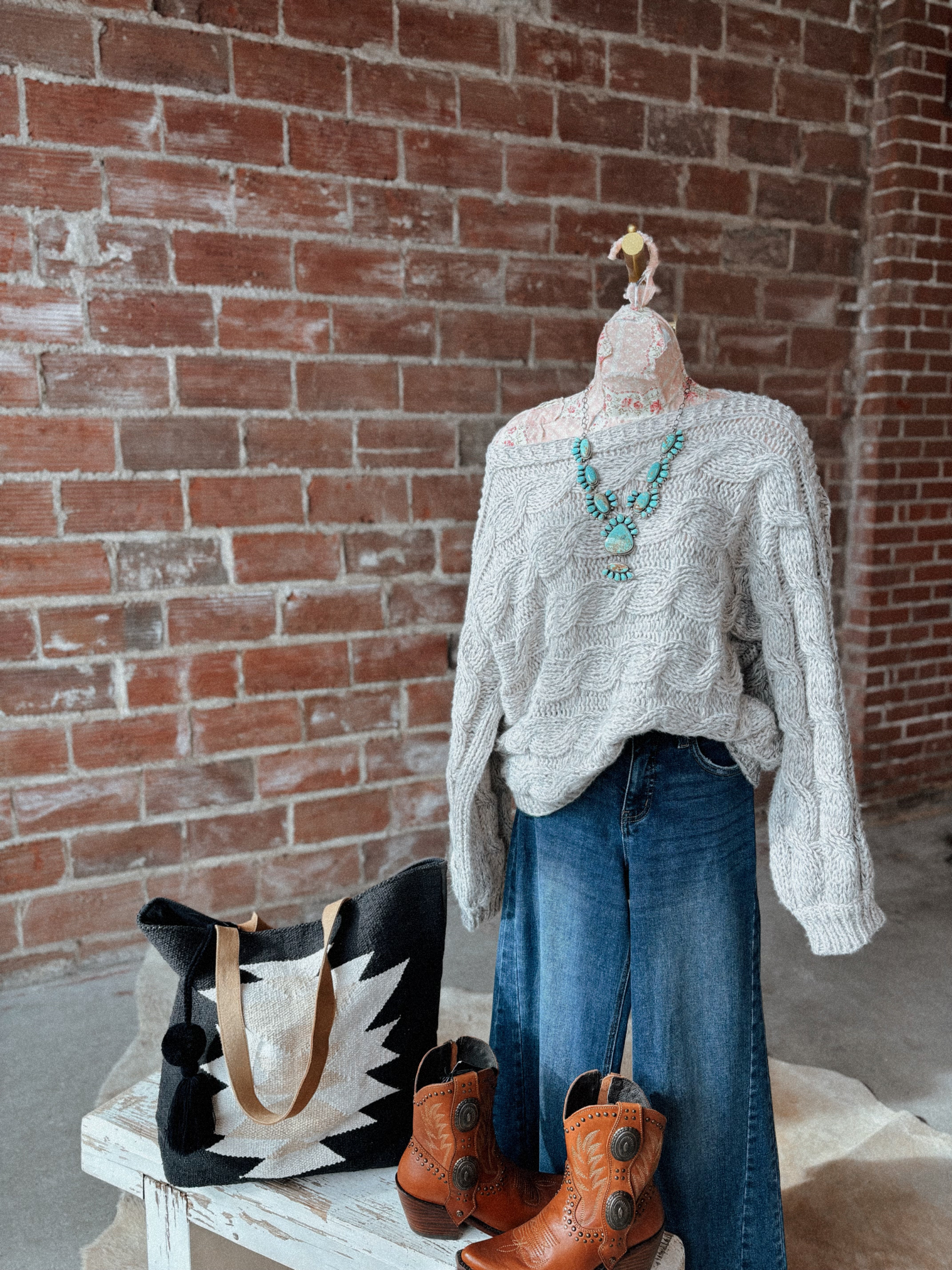 Mannequin wearing a white sweater and blue jeans with a black and white bag and brown boots against a brick wall.
