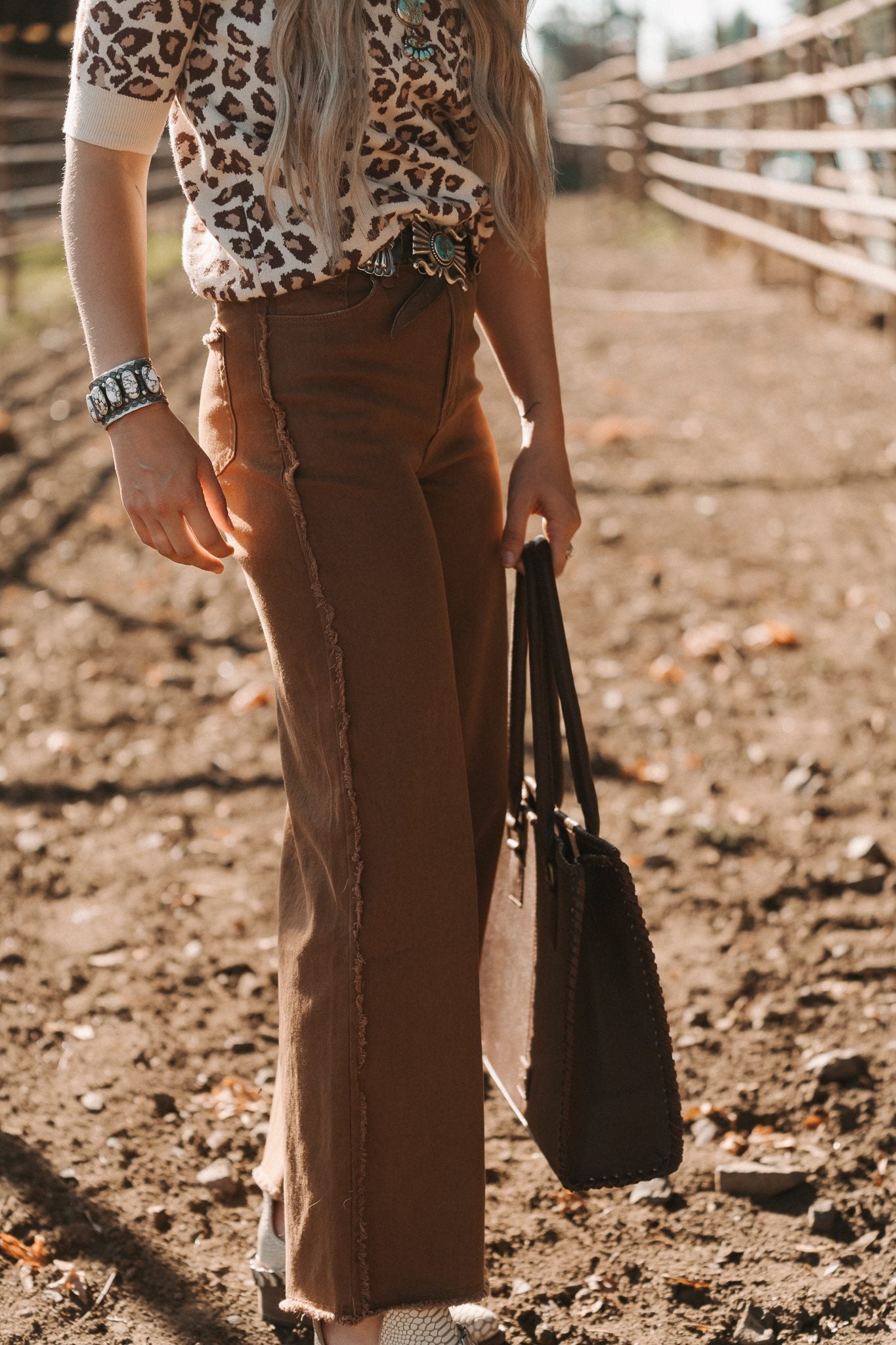 Person wearing a leopard print top and brown pants holding a black handbag on a dirt path.