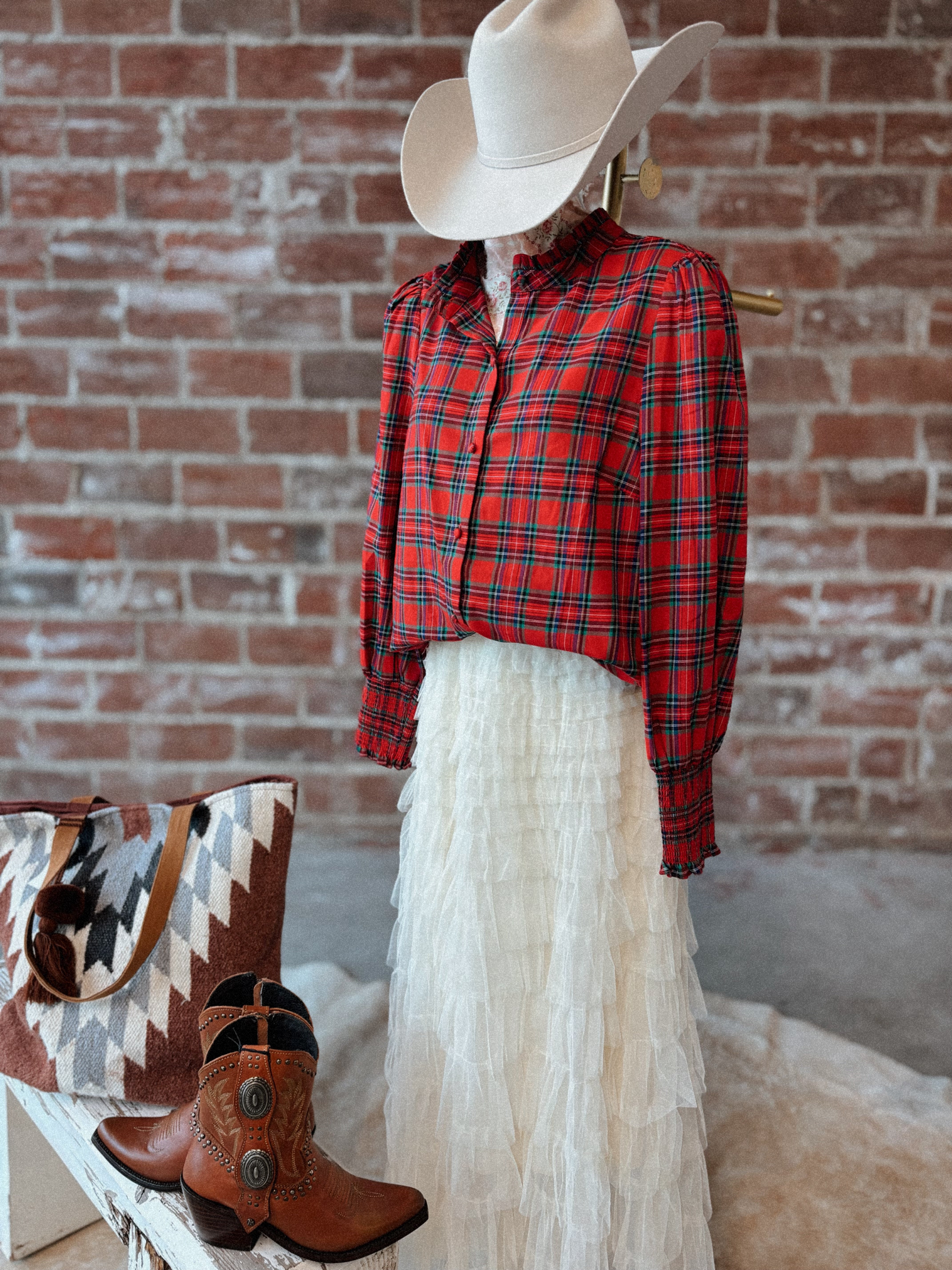 Person wearing a red plaid shirt, white skirt, cowboy hat, and boots with a patterned bag against a brick wall.