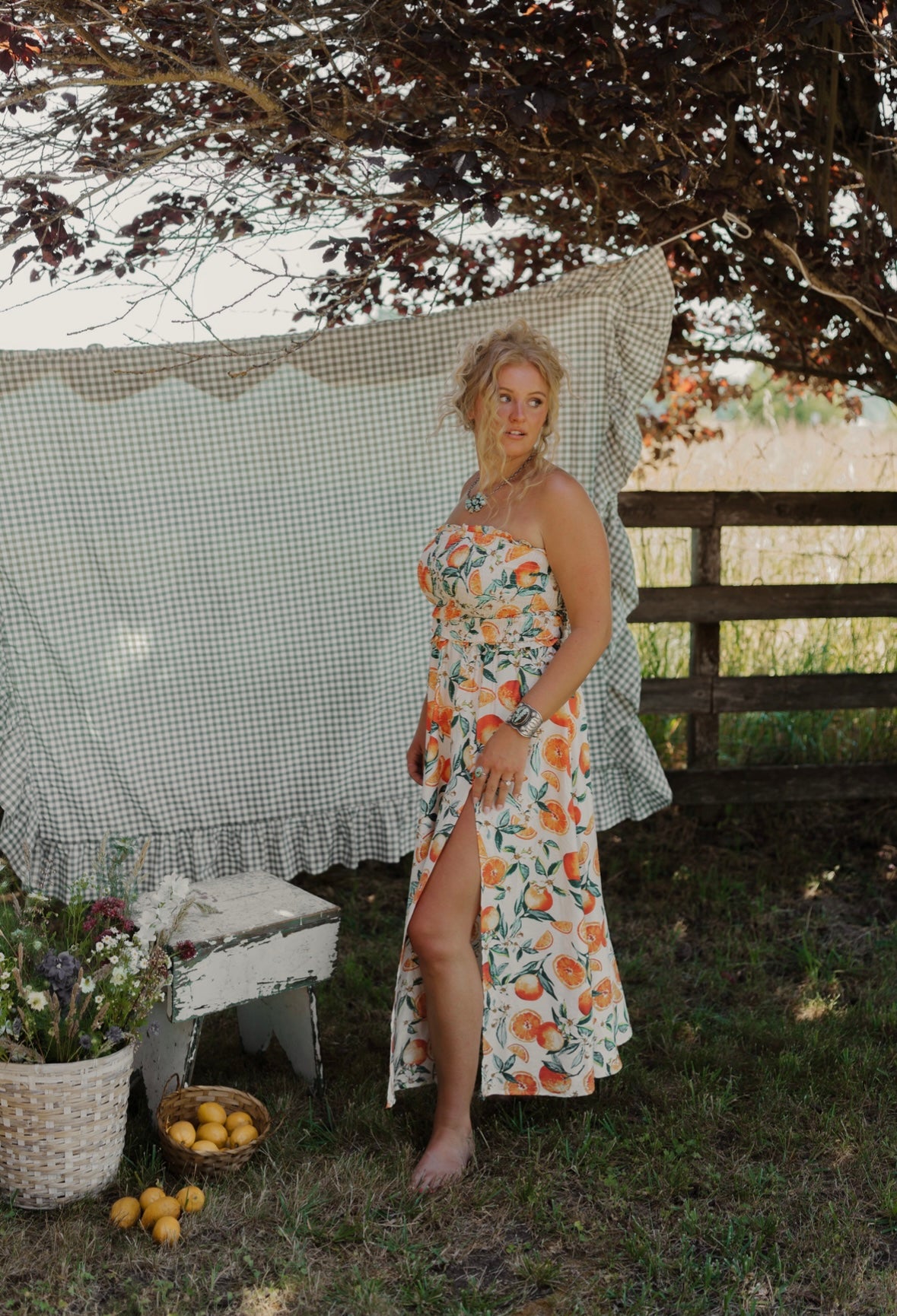 Woman in a floral dress standing outdoors with a checkered curtain and wooden fence in the background.