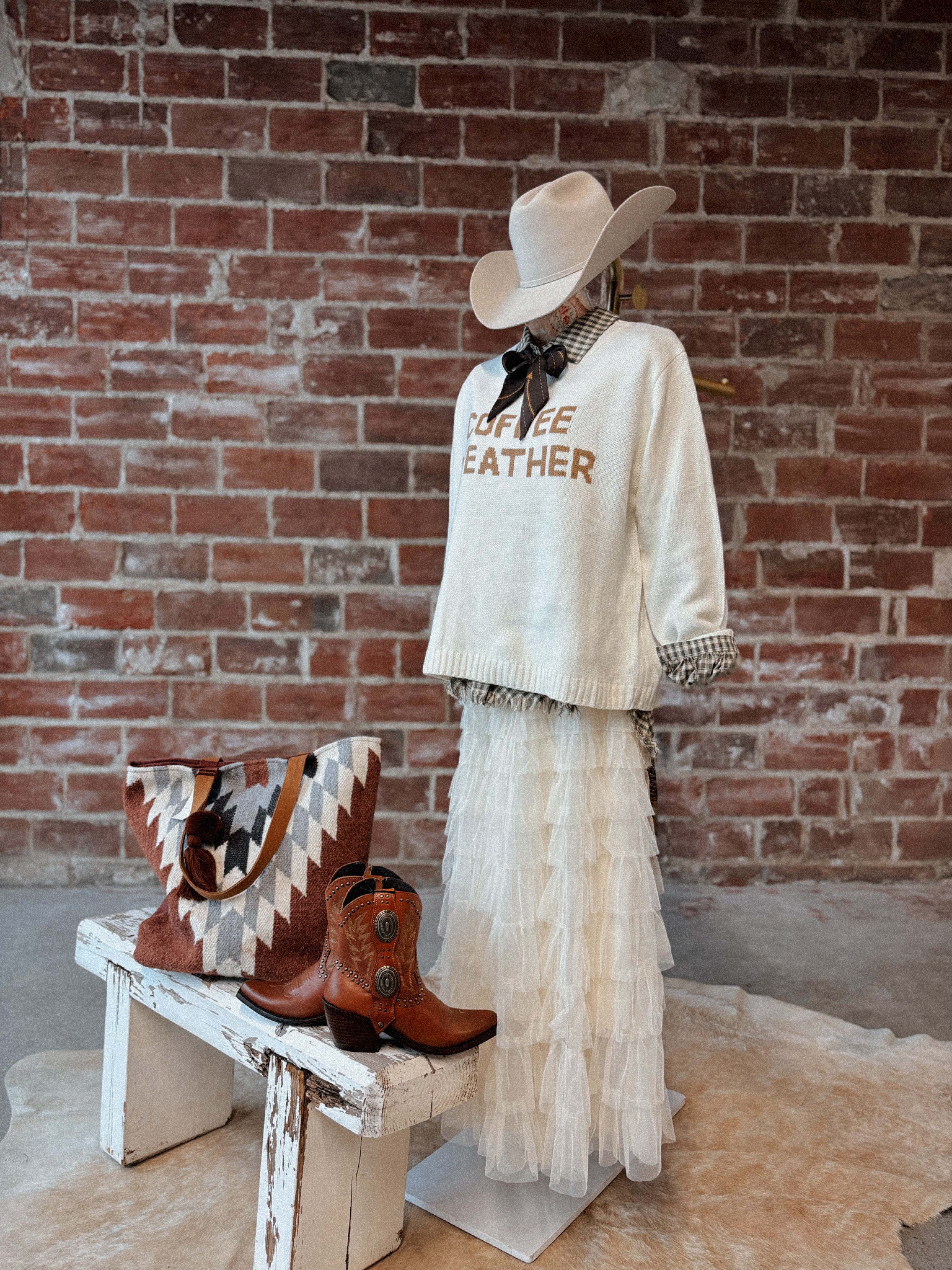 Mannequin wearing a white sweater with text, white skirt, and cowboy hat against a brick wall.