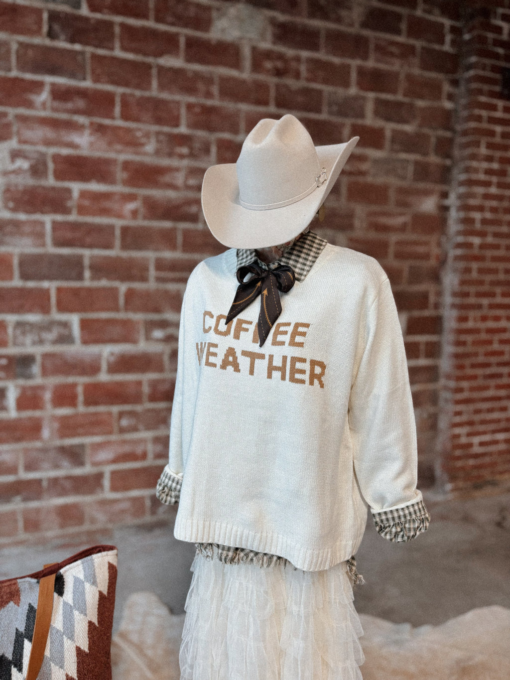 Person wearing a white sweater with 'Coffee Weather' text, white skirt, and cowboy hat against a brick wall.