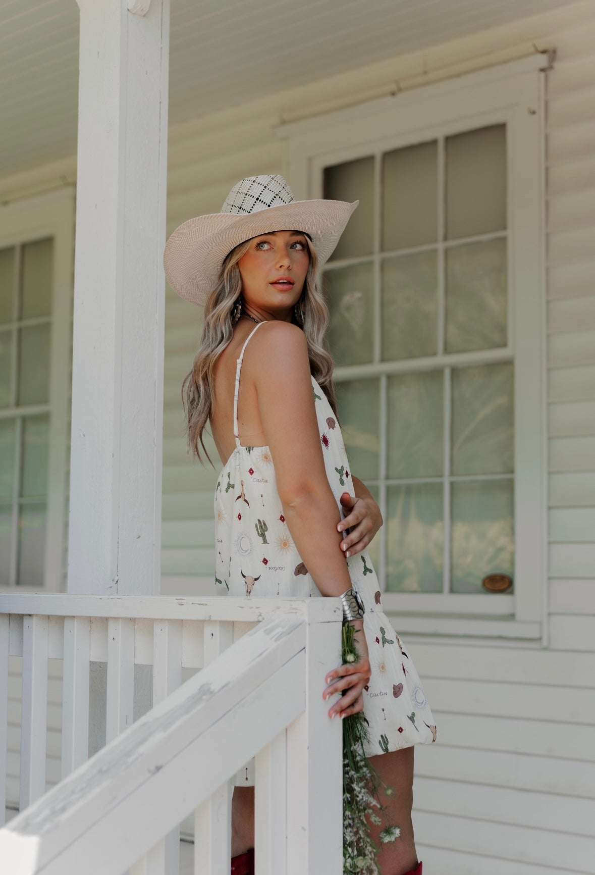 Woman in a white dress with floral patterns and a wide-brimmed hat standing on a porch.