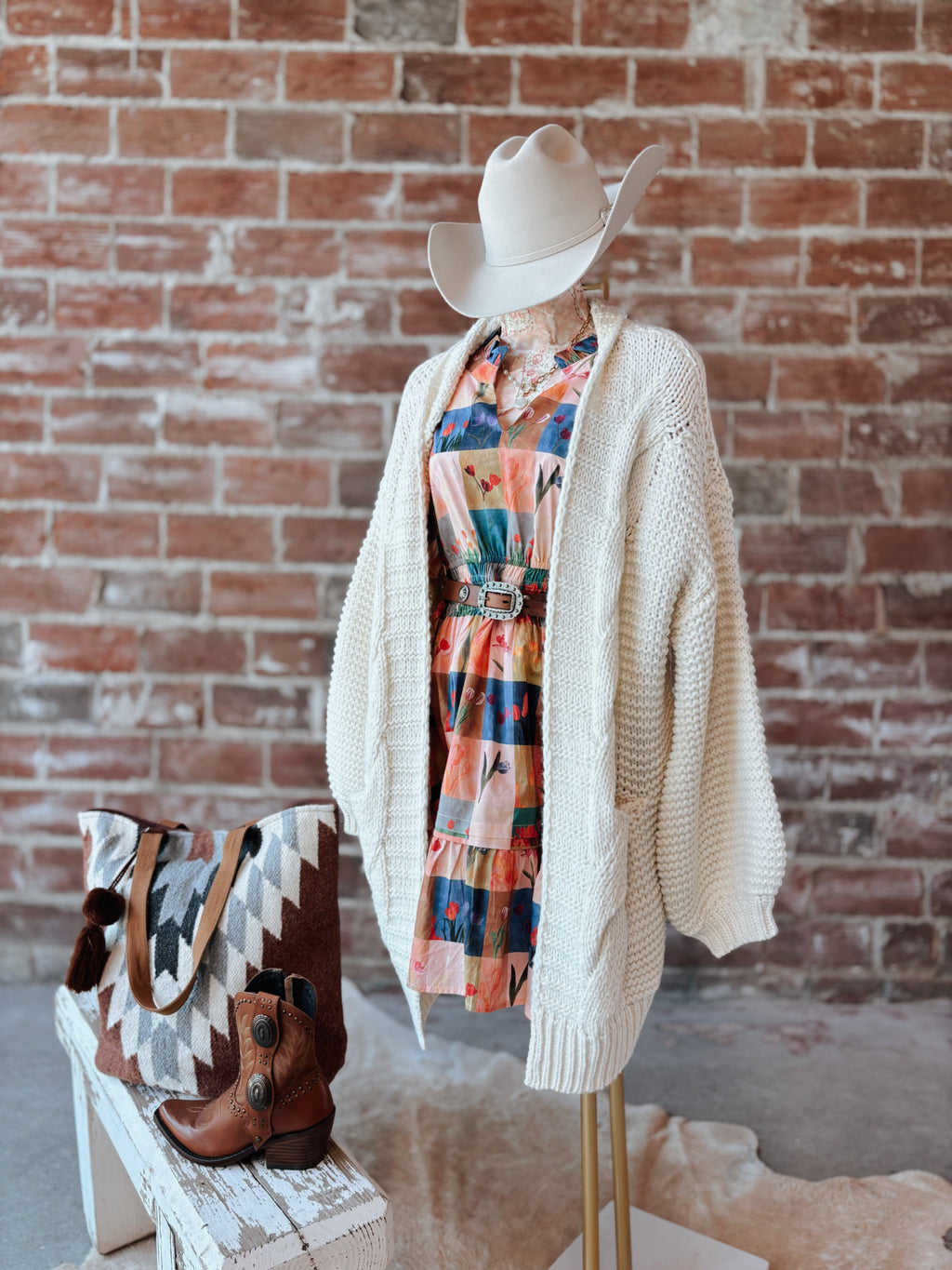 Mannequin wearing a beige cardigan, colorful scarf, and cowboy hat against a brick wall.