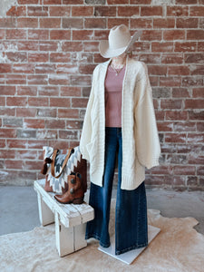 Person wearing a beige cardigan, pink top, blue jeans, and cowboy hat against a brick wall.