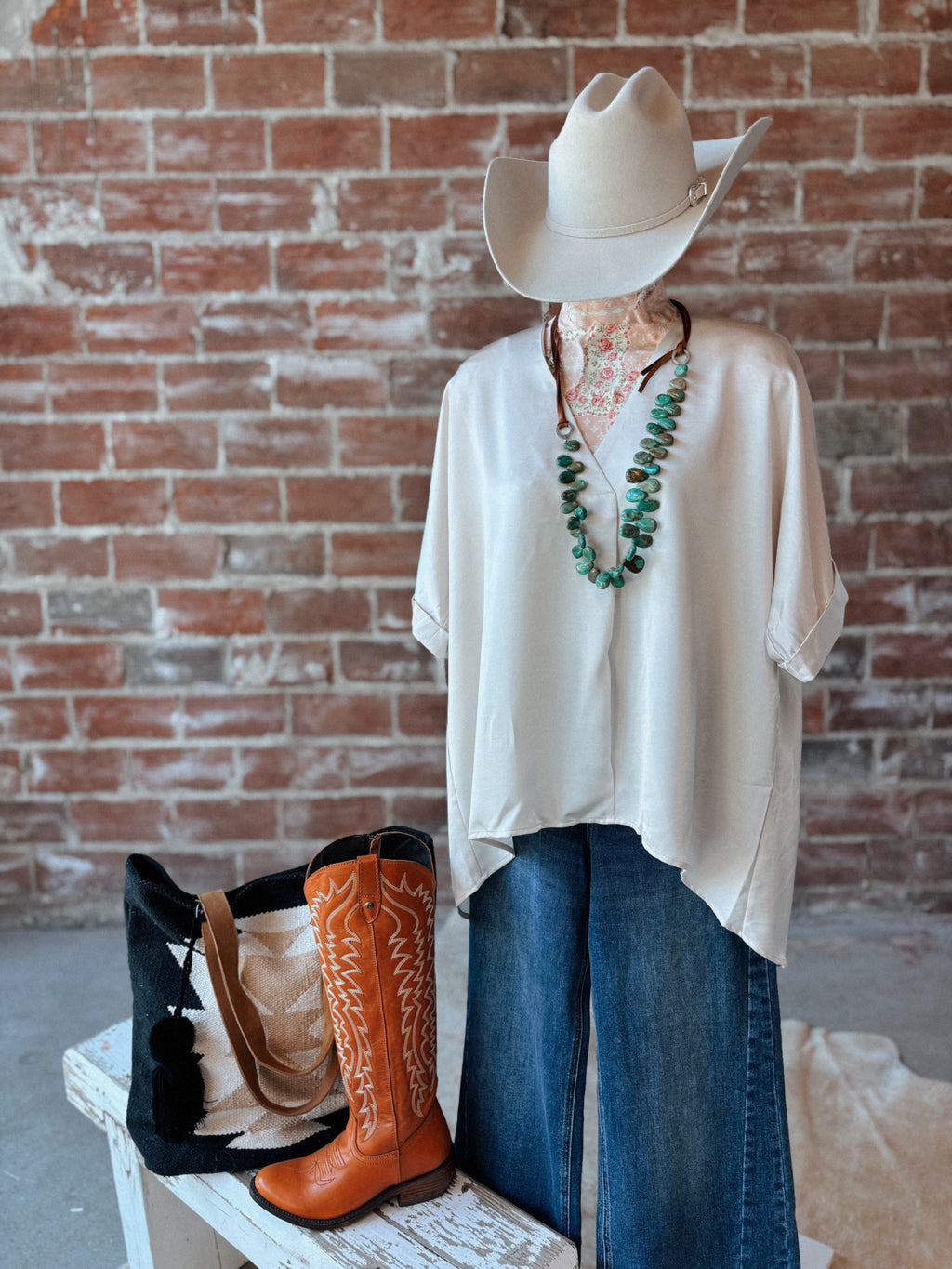 Mannequin wearing a white shirt, blue jeans, and cowboy boots with a cowboy hat against a brick wall.