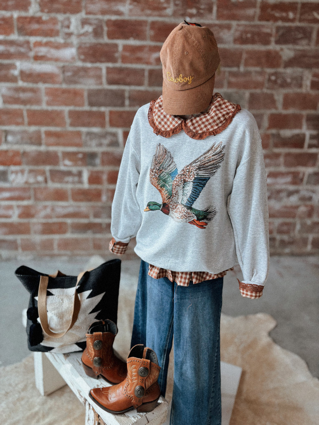 Person wearing a sweater with a bird design, plaid shirt, and jeans against a brick wall.