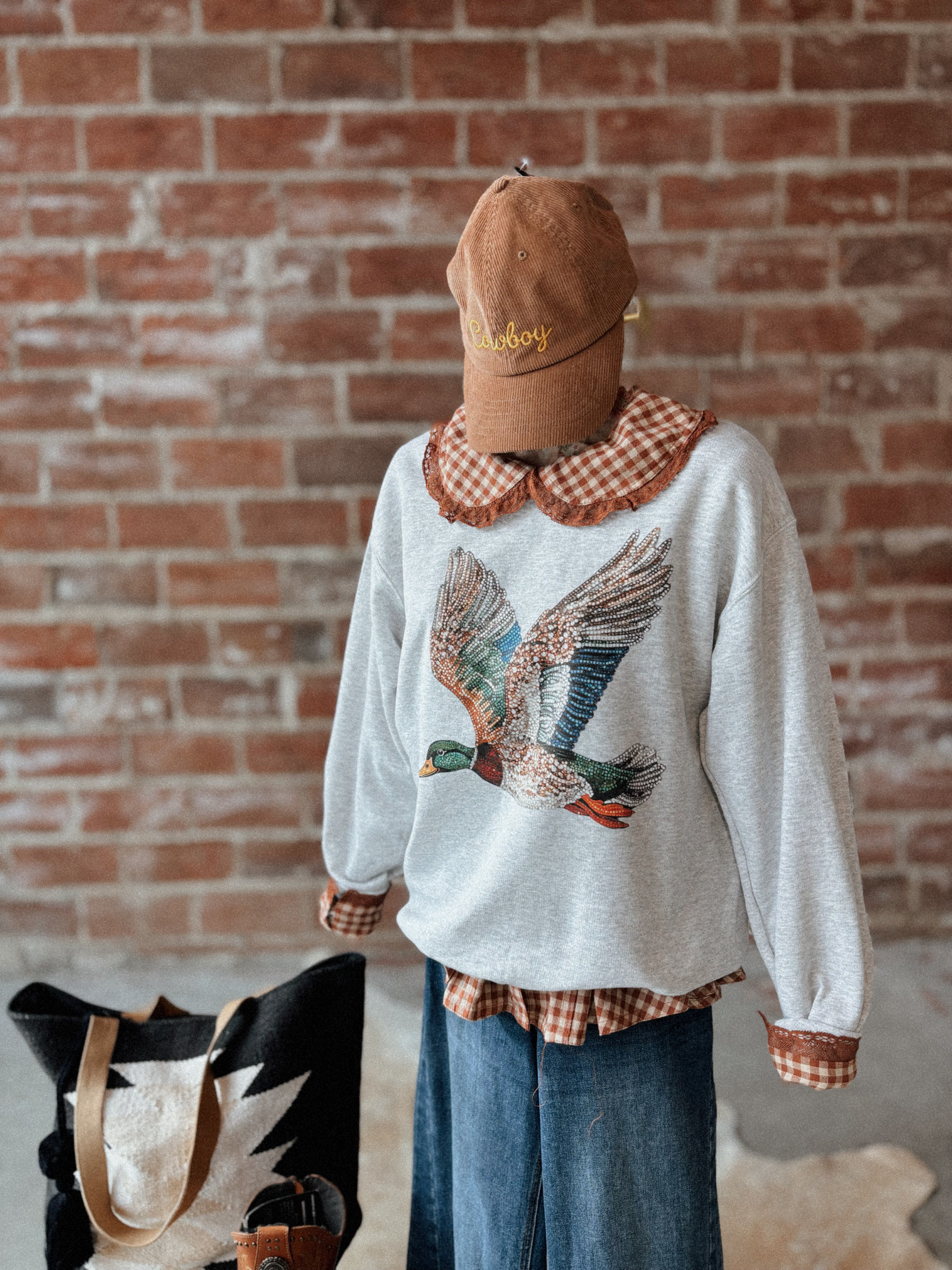 Person wearing a sweater with a duck design, brown cap, and plaid shirt against a brick wall.