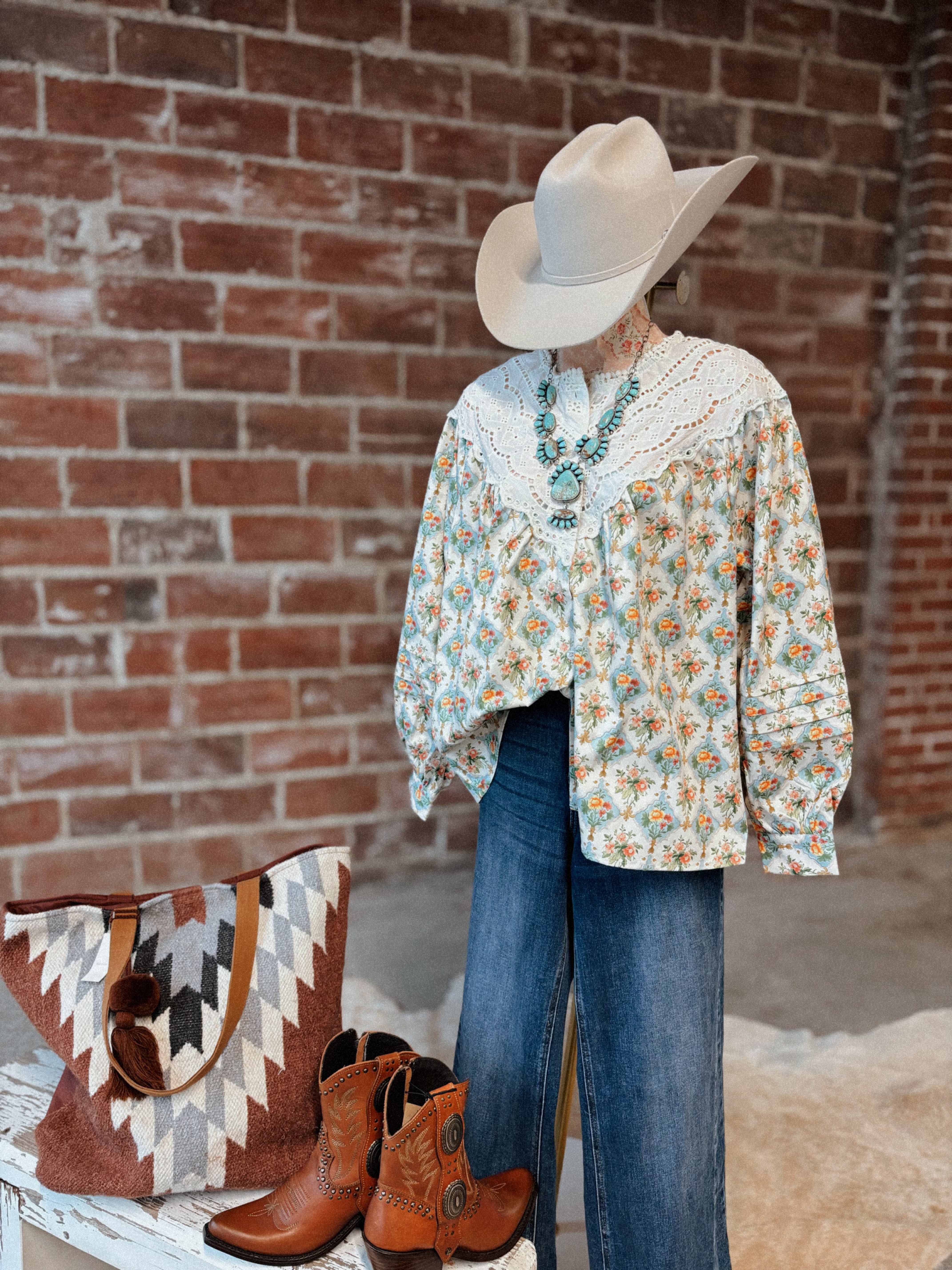 Person wearing a floral blouse, jeans, cowboy hat, and boots with a patterned bag against a brick wall.