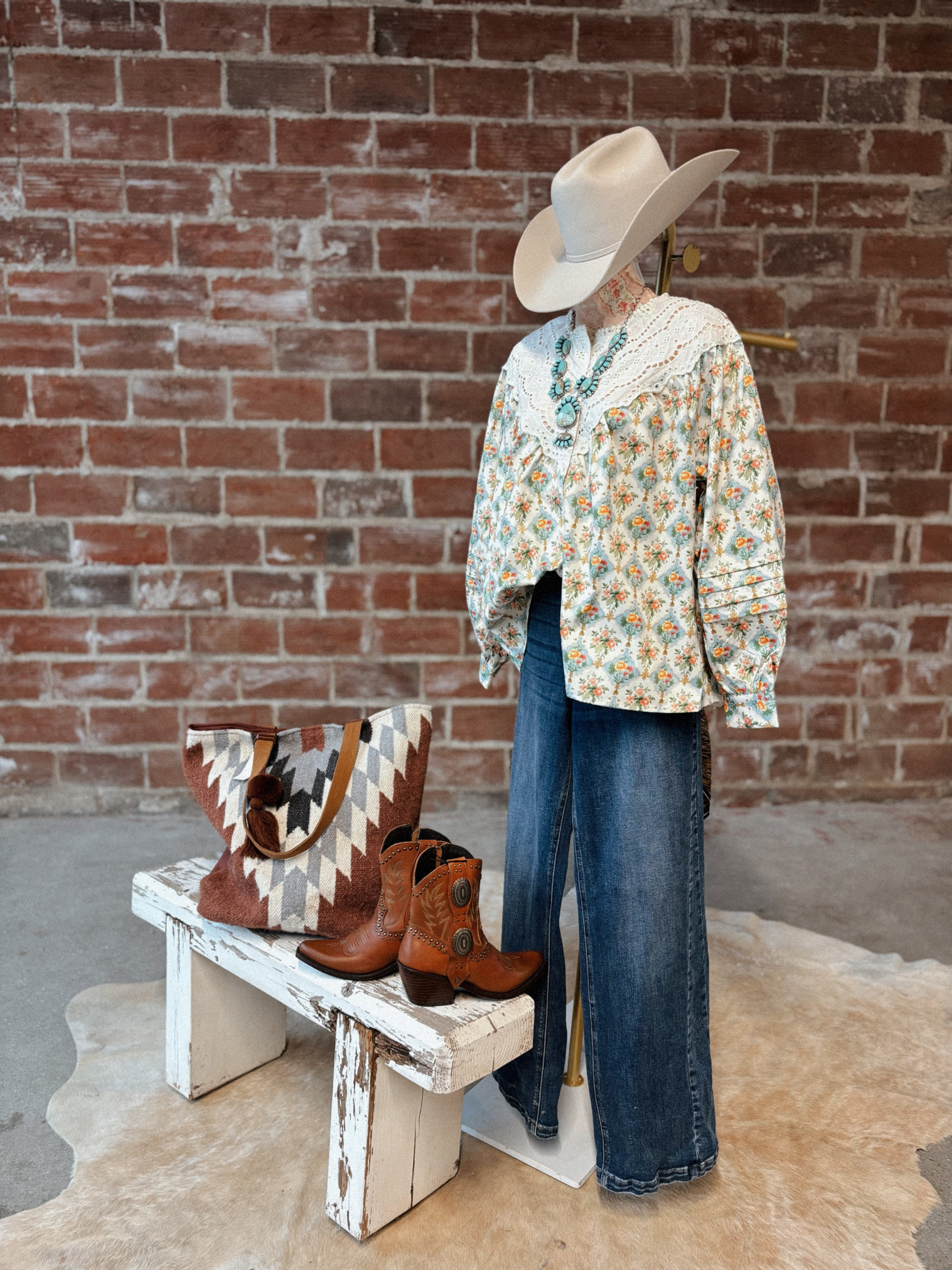 Mannequin wearing a floral blouse, jeans, and cowboy boots with a cowhide rug and bench in front of a brick wall.