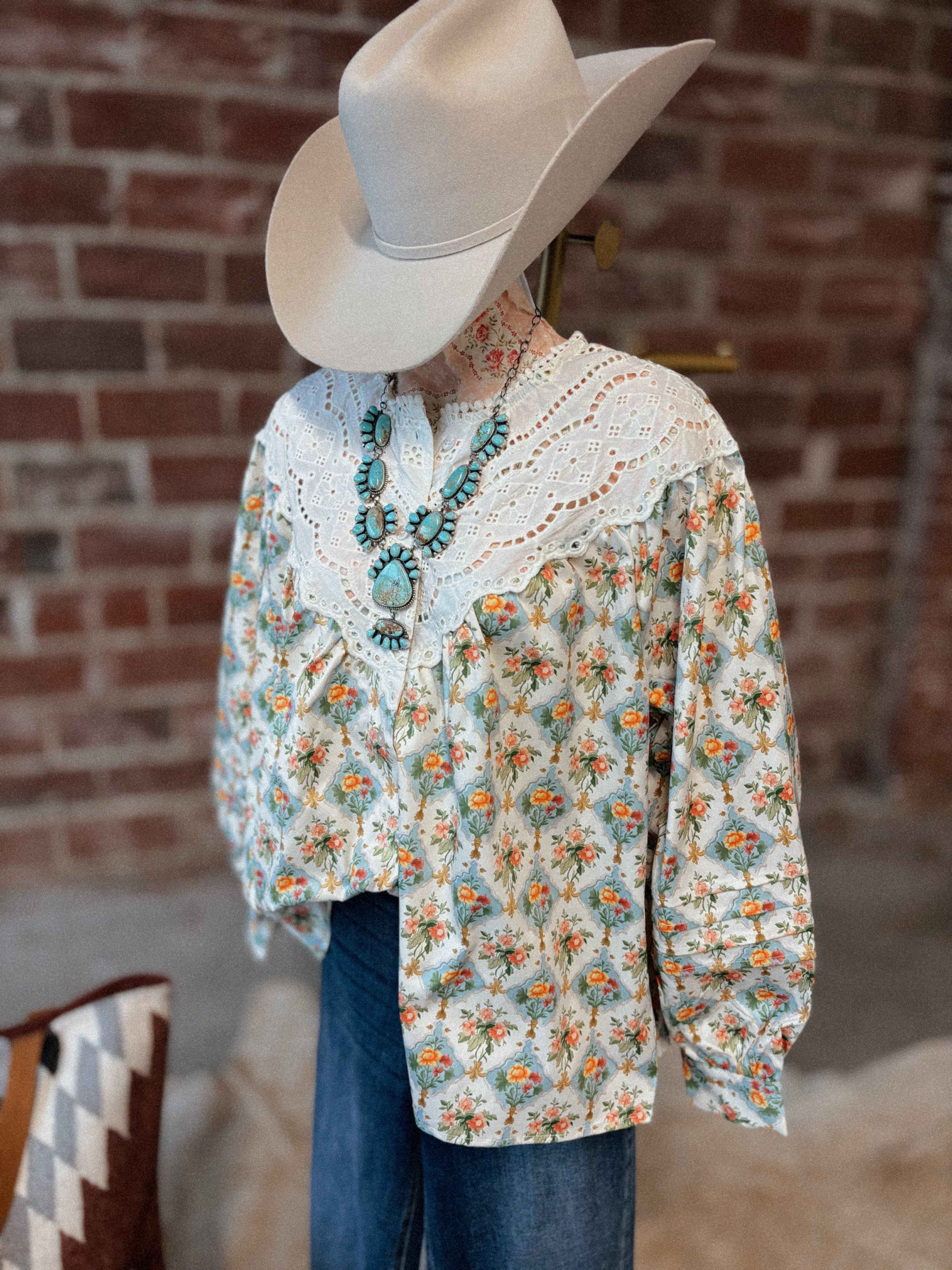 Person wearing a floral blouse, cowboy hat, and blue jeans against a brick wall.