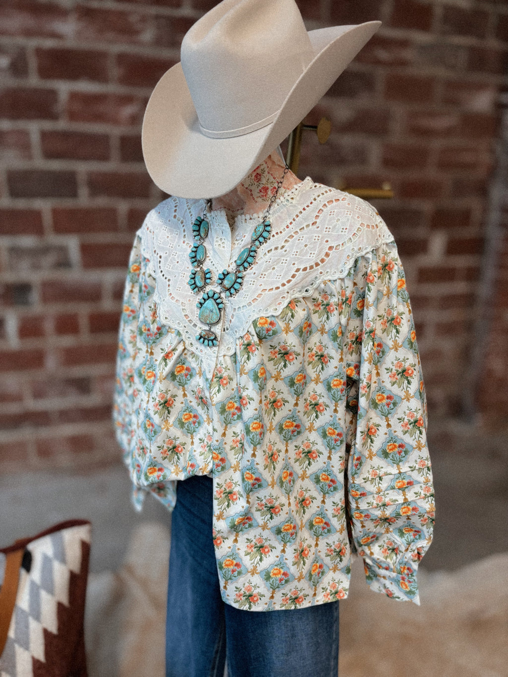 Person wearing a floral blouse, cowboy hat, and blue jeans against a brick wall.