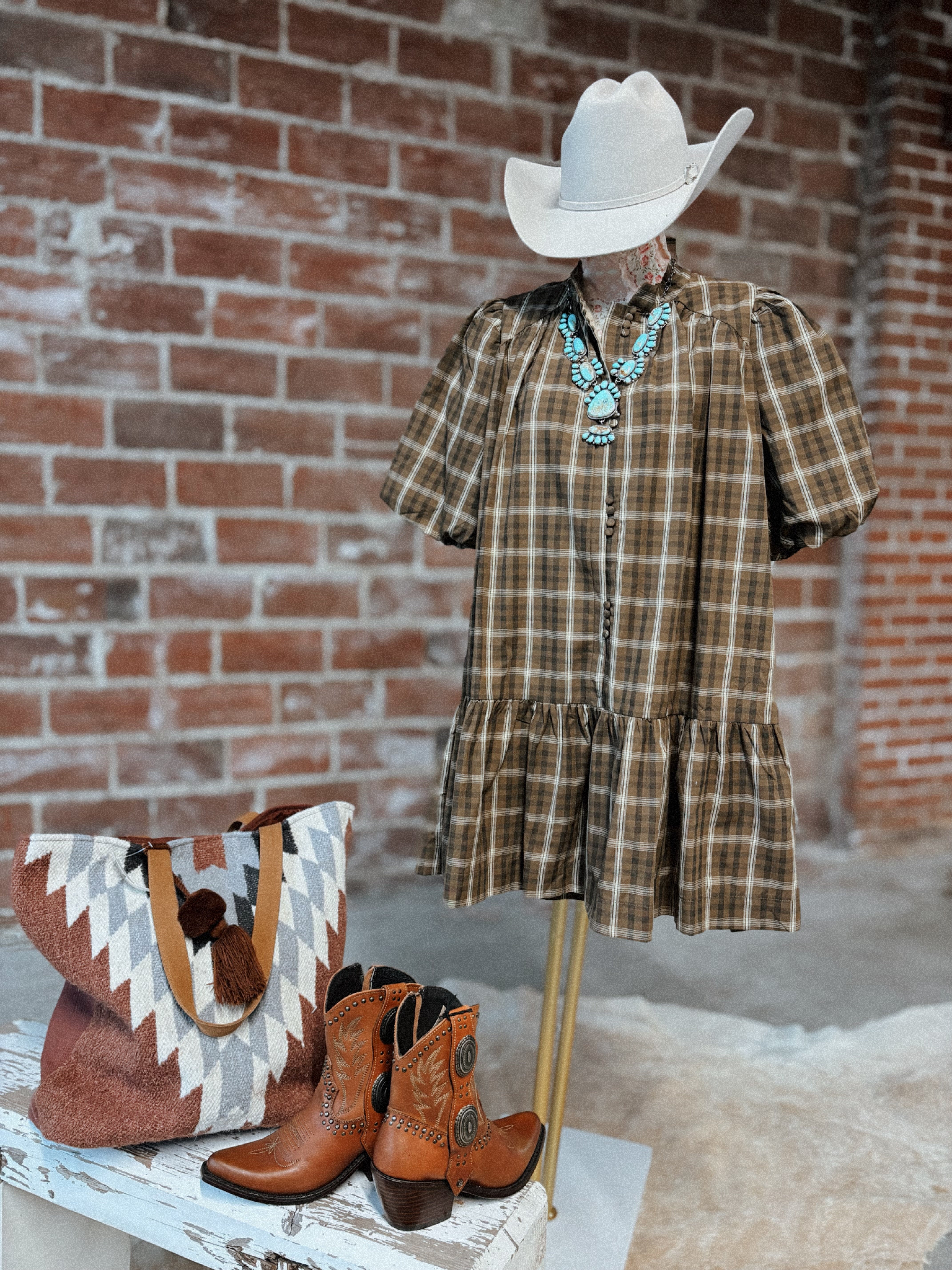 Mannequin wearing a plaid dress with cowboy hat, boots, and bag against a brick wall.