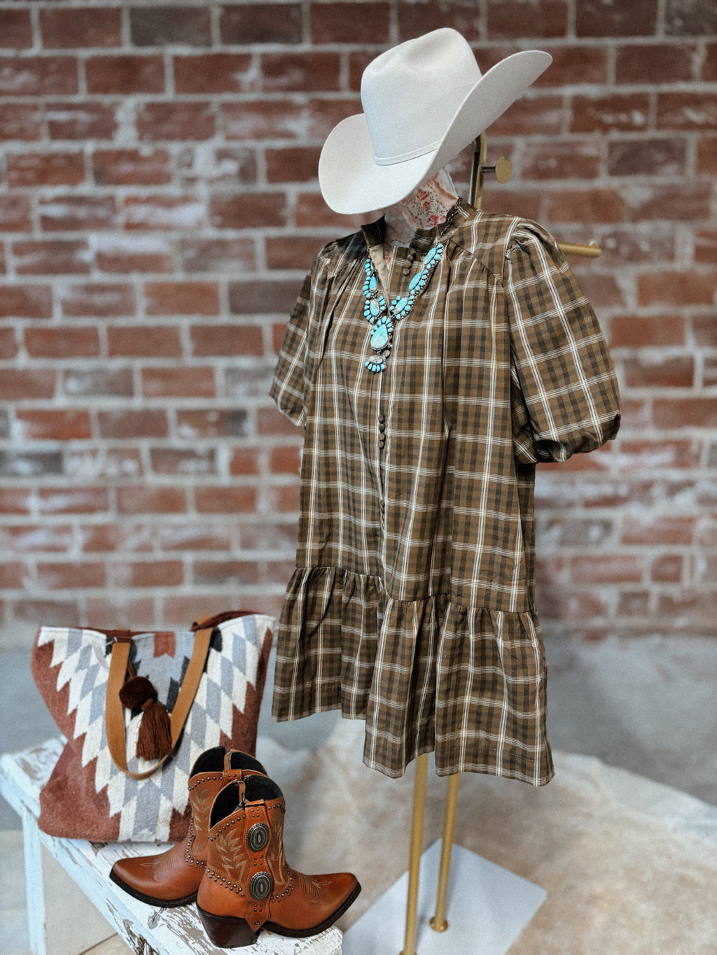 Mannequin wearing a plaid dress, cowboy hat, and boots with a handbag against a brick wall.