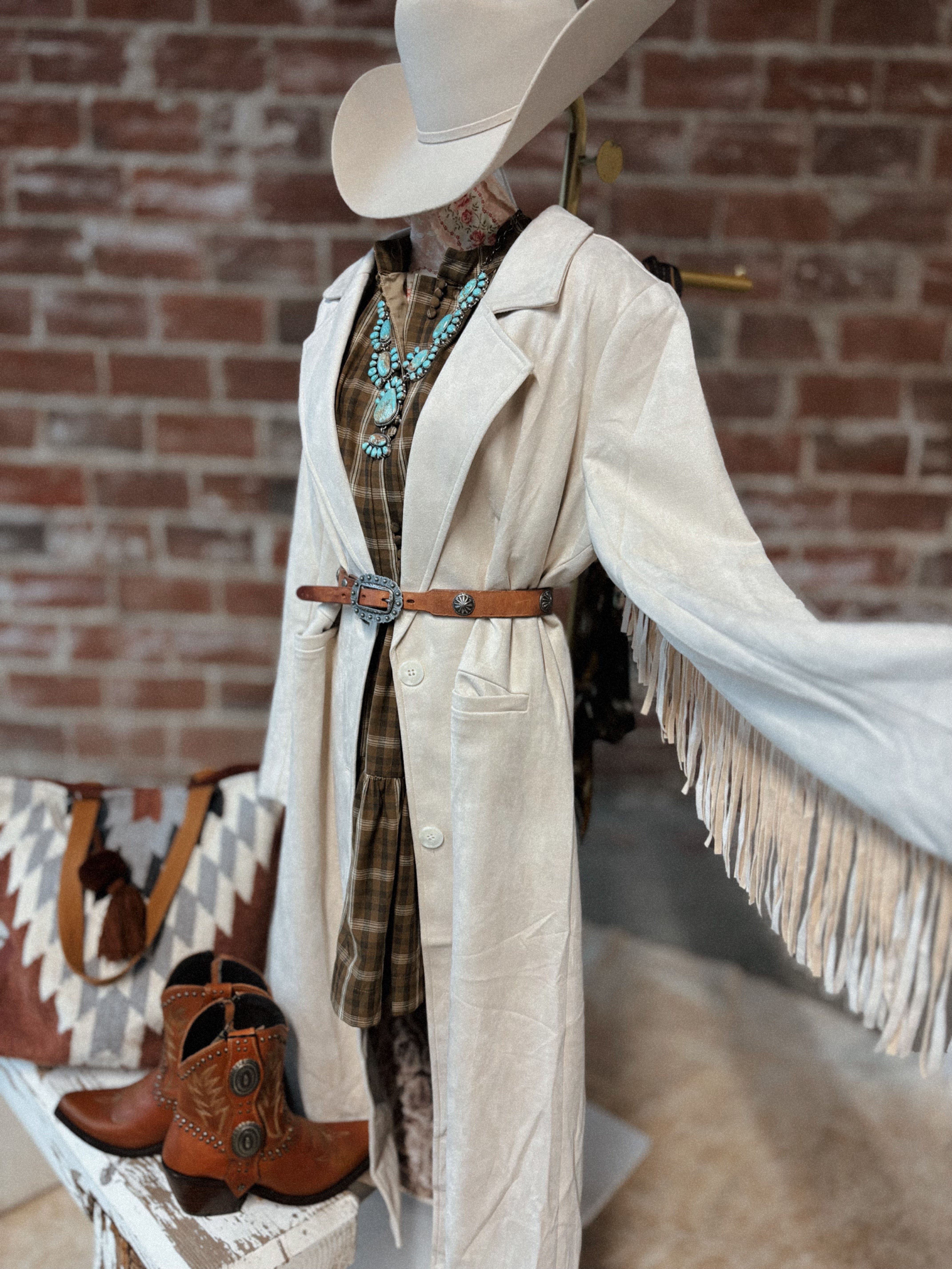 Person wearing a long beige coat with fringe, plaid shirt, and cowboy boots against a brick wall.