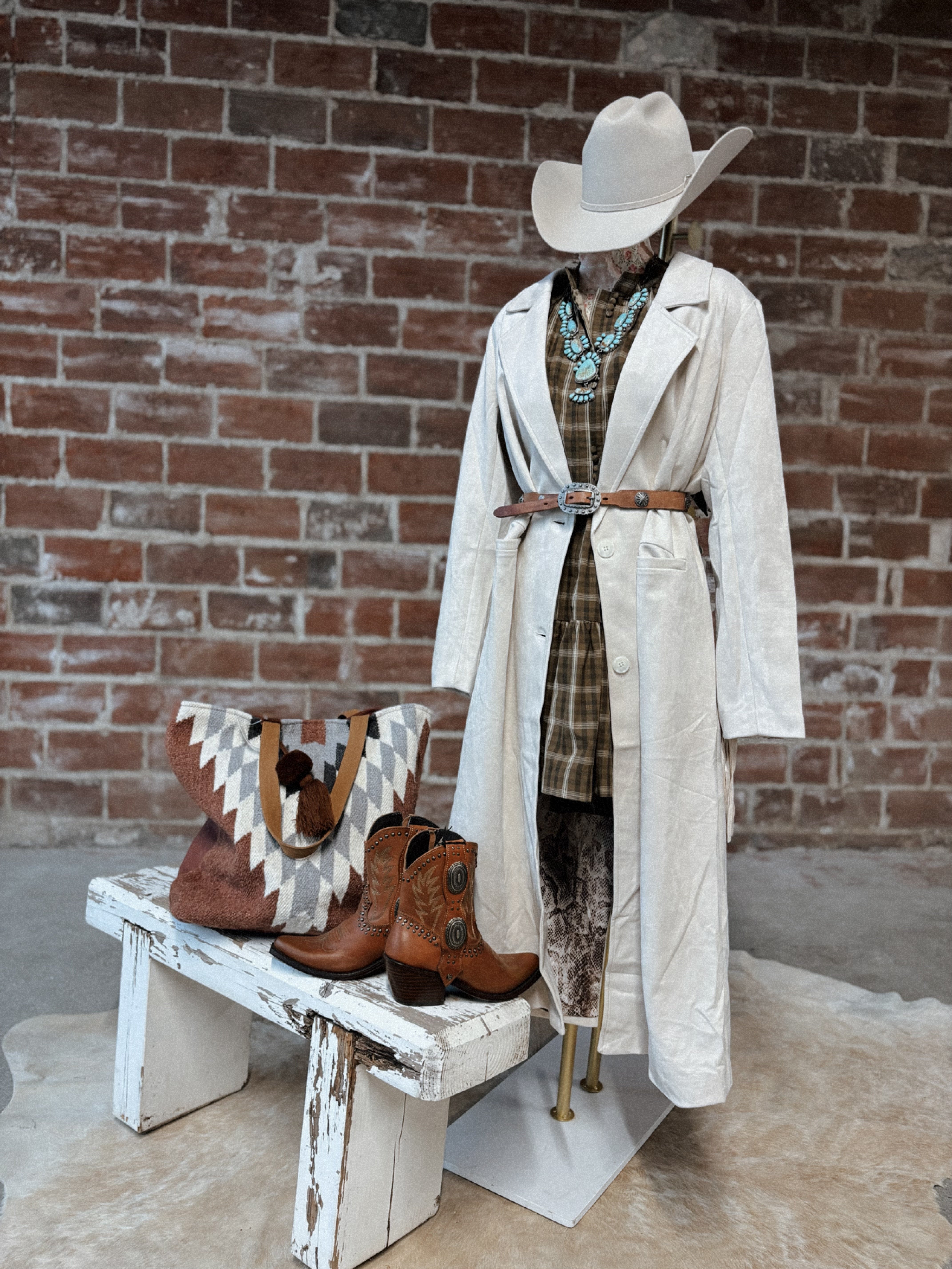Mannequin wearing a white coat and cowboy hat with boots on a bench against a brick wall.