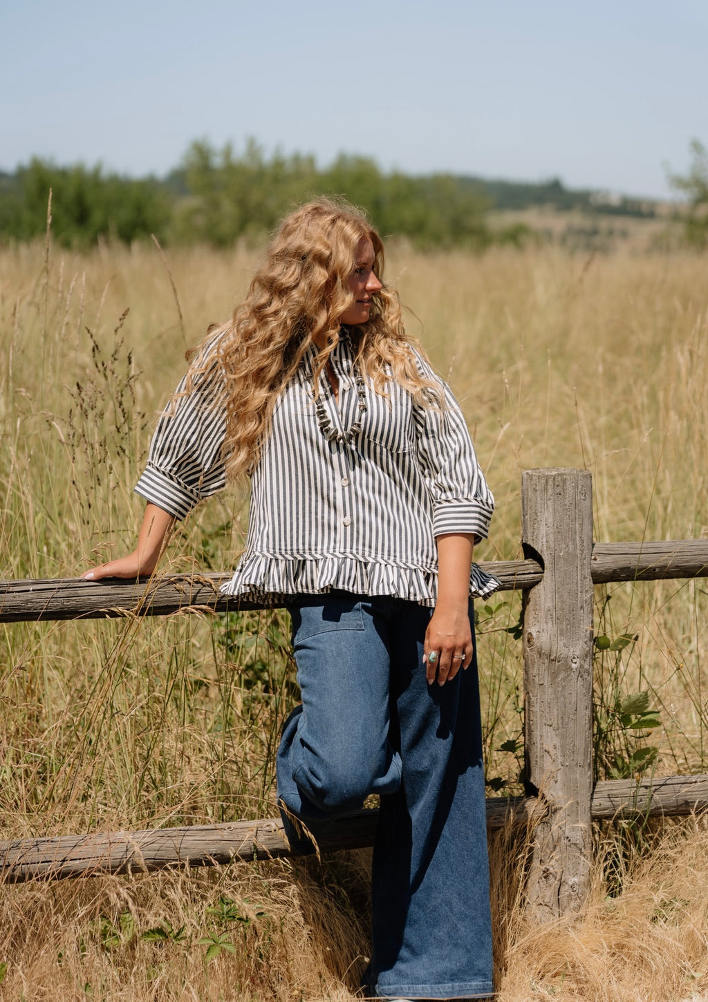 Woman leaning on a wooden fence in a field with tall grass and trees in the background