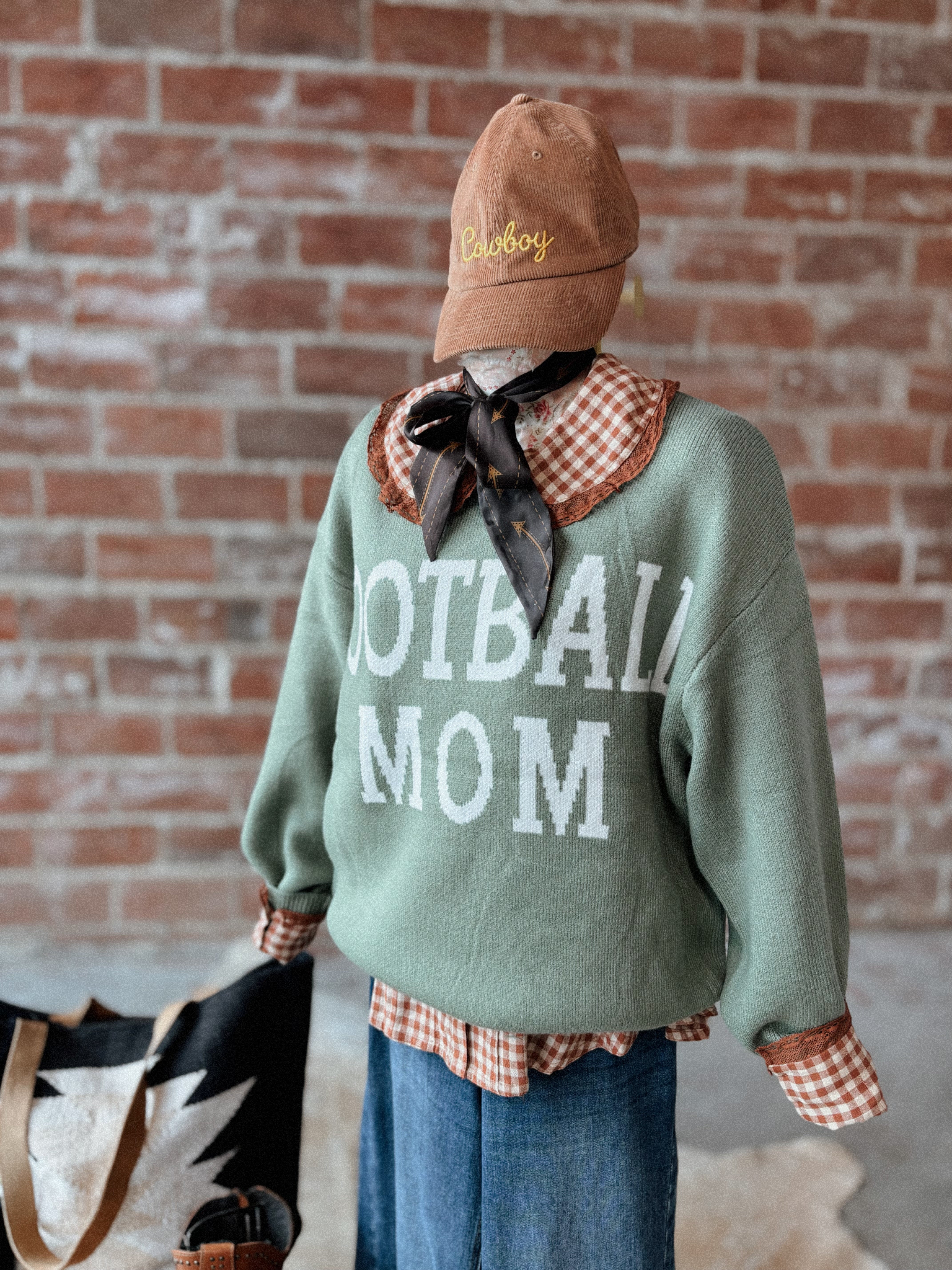 Person wearing a 'Football Mom' sweater with a cowboy hat and scarf, standing against a brick wall.