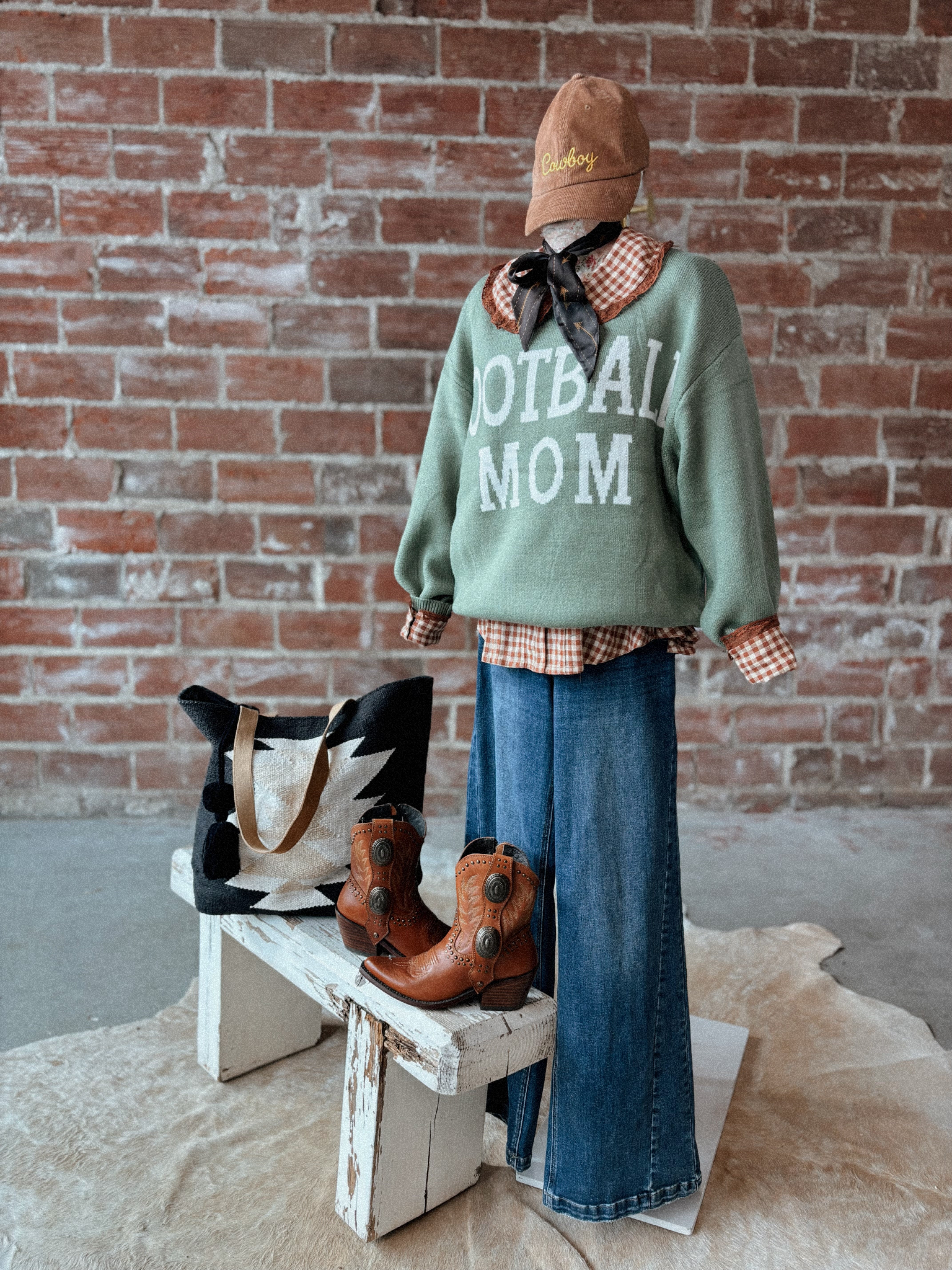 Person wearing a 'Football Mom' sweater with boots and a hat, standing against a brick wall.