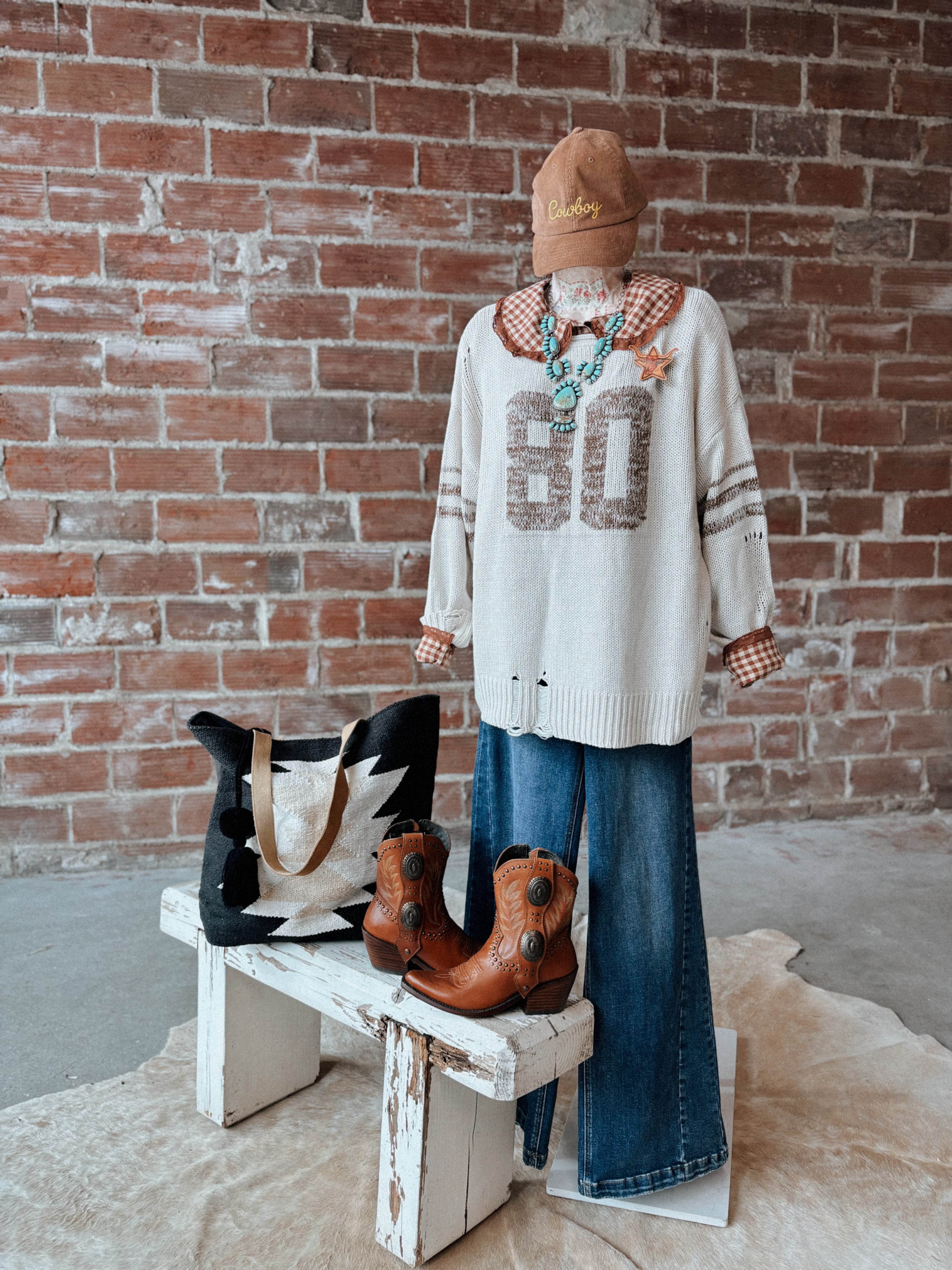 Mannequin wearing a sweater with 'BOO' text, jeans, and boots against a brick wall.