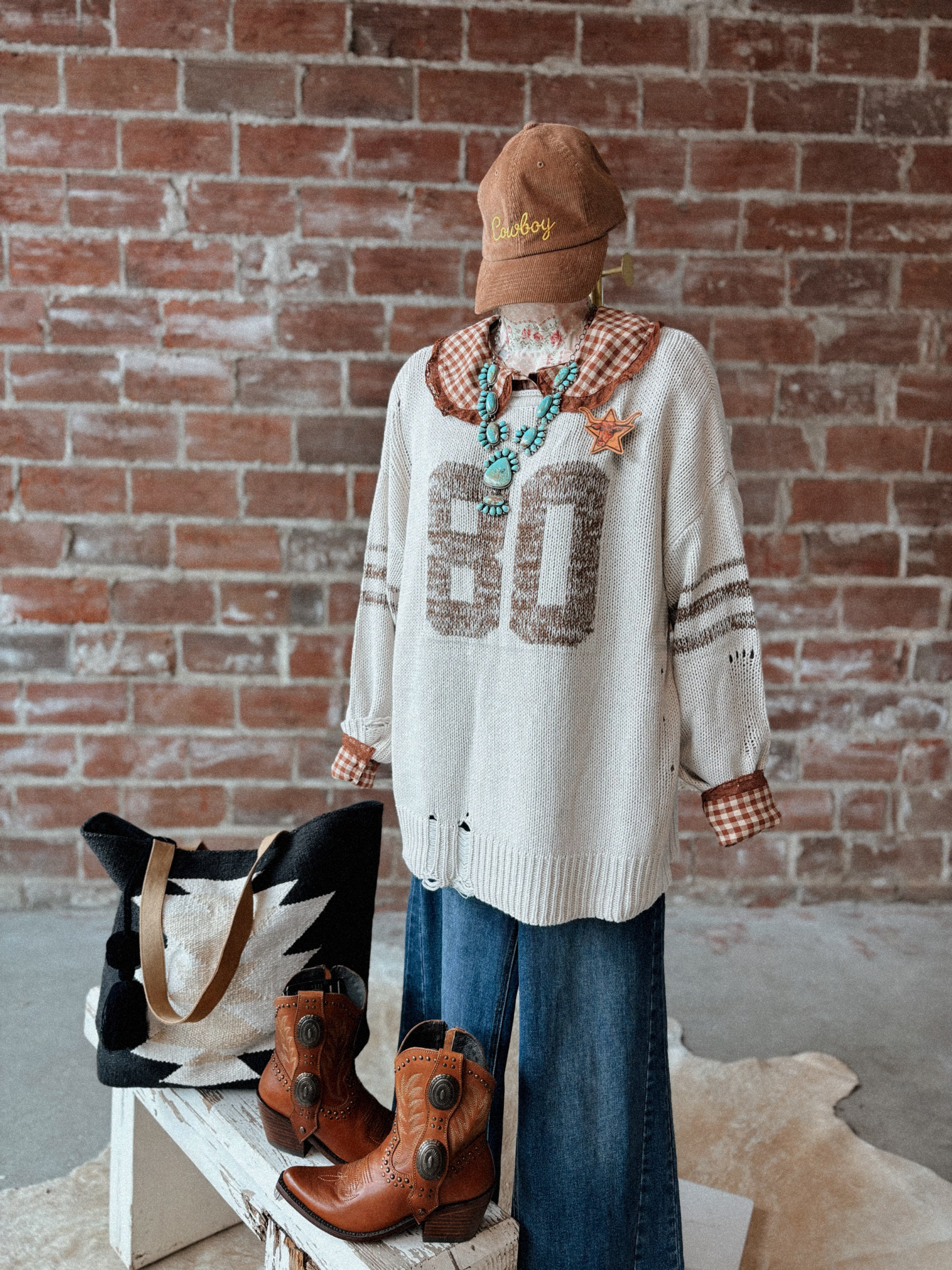 Person wearing a patterned sweater and jeans with boots and a bag on a bench against a brick wall.