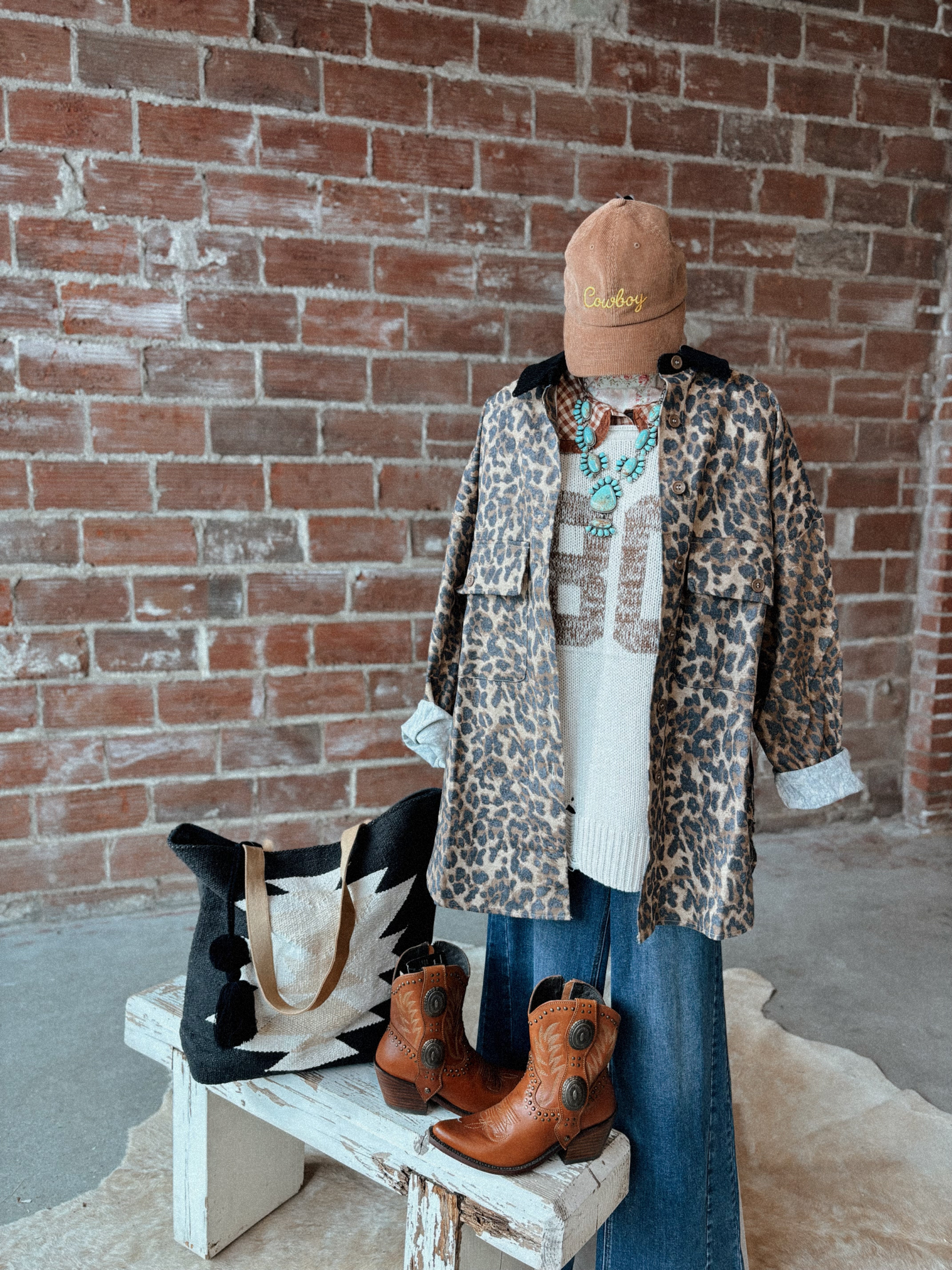 Person wearing a leopard print coat, brown boots, and a beige cap against a brick wall.