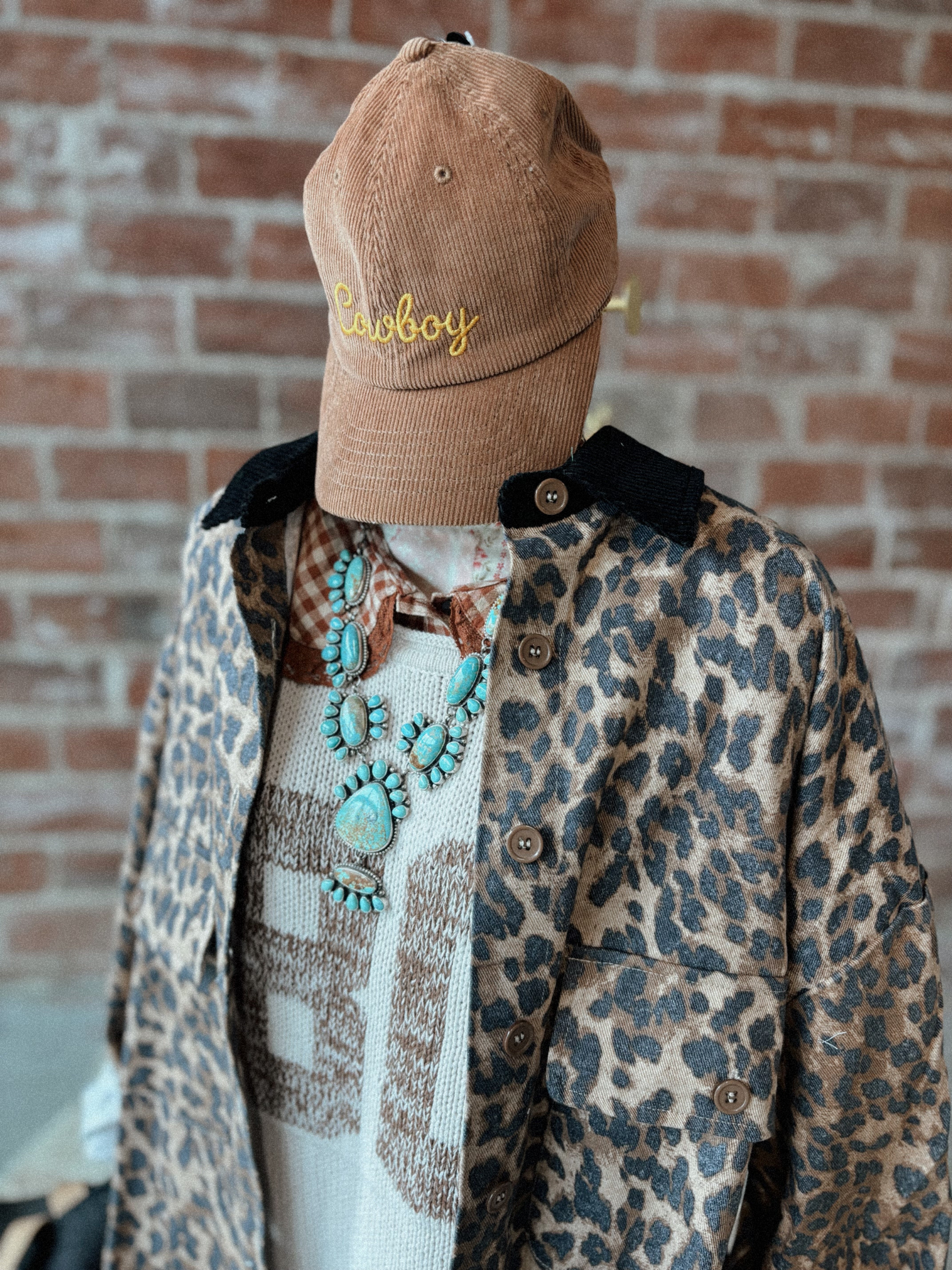 Mannequin wearing a leopard print coat, brown cap with 'Cowboy' text, and turquoise necklace against a brick wall.