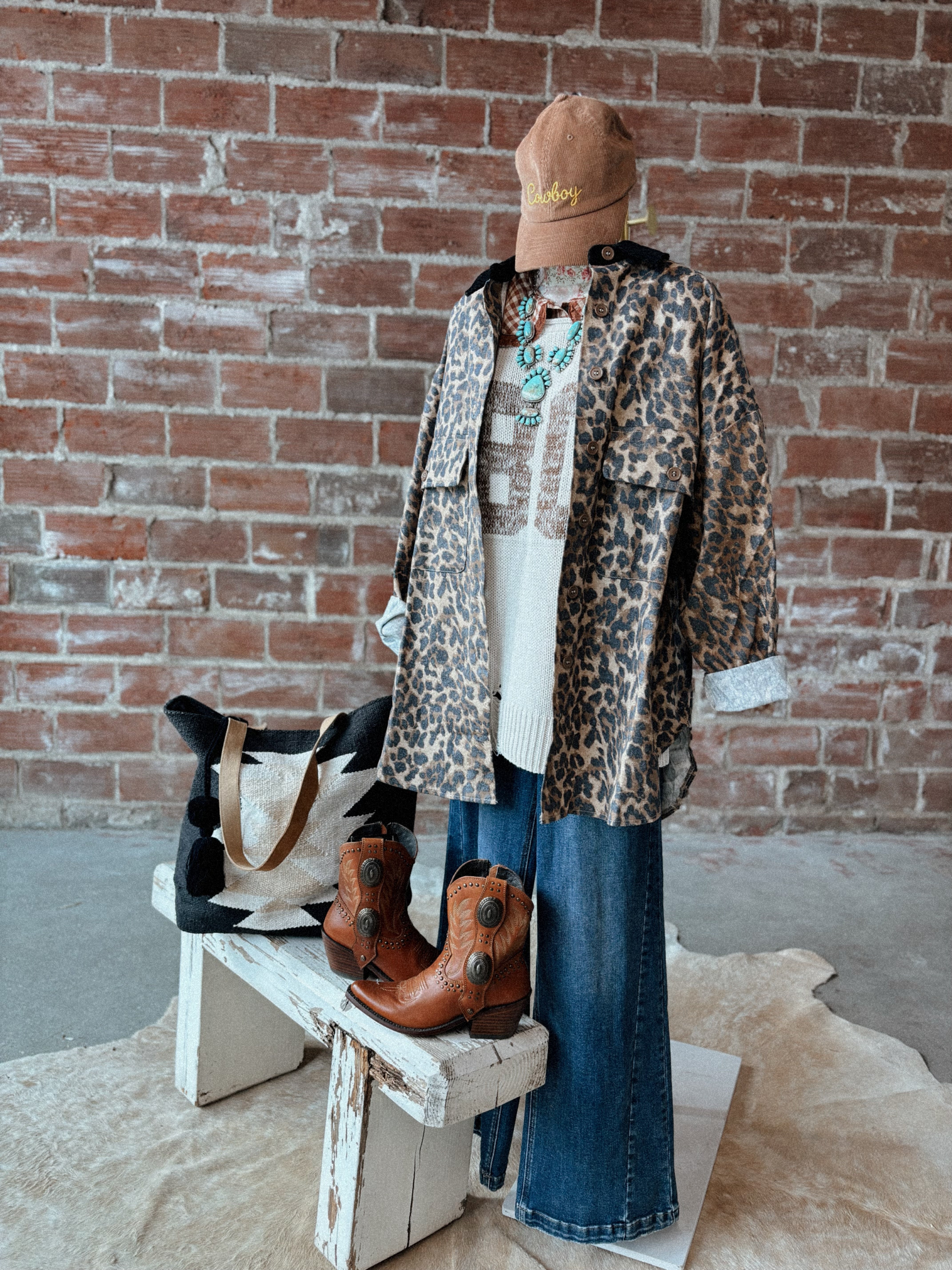 Person wearing a leopard print coat, blue jeans, and brown boots against a brick wall.