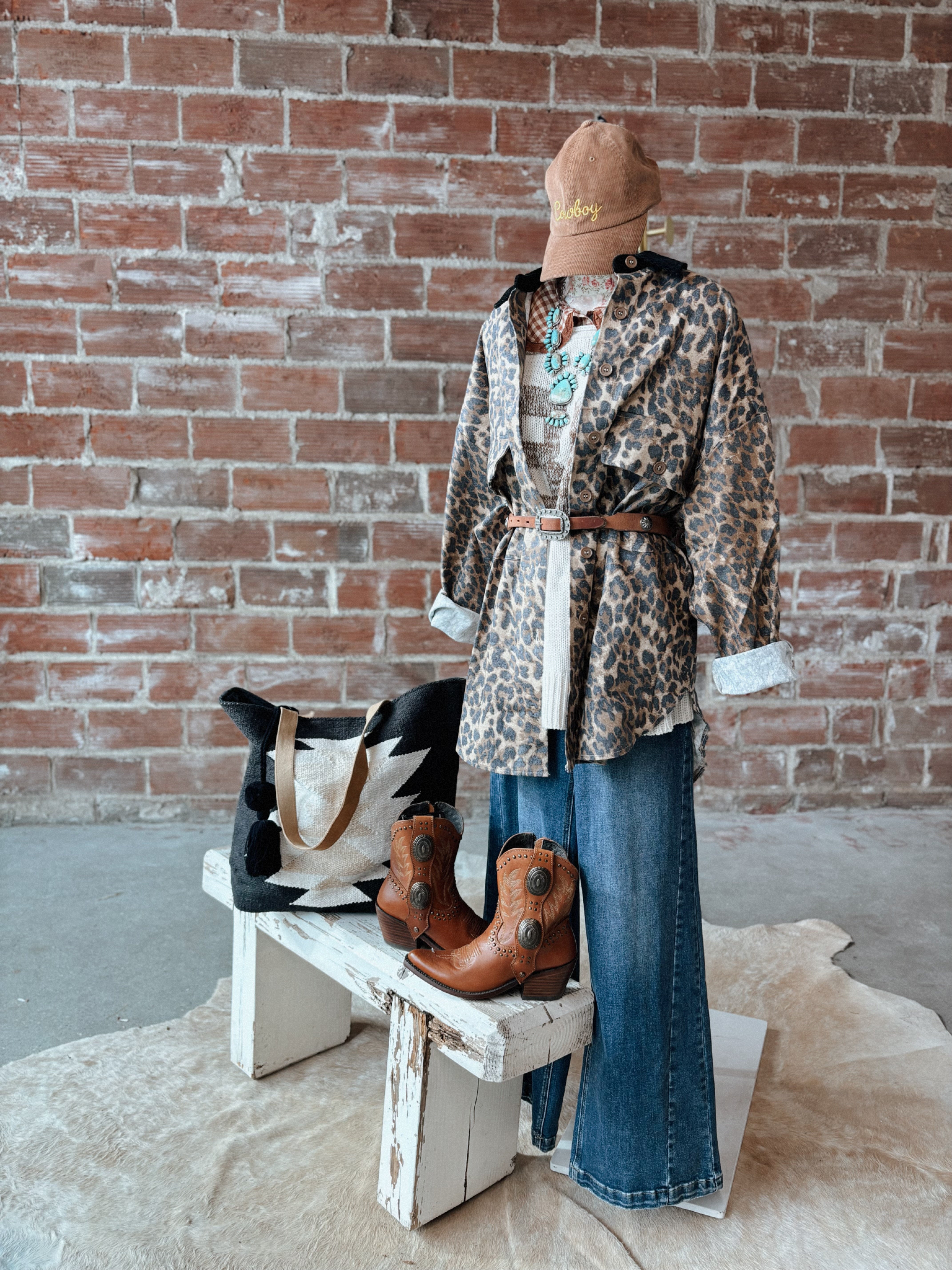 Mannequin wearing a leopard print jacket, blue jeans, and brown boots against a brick wall.
