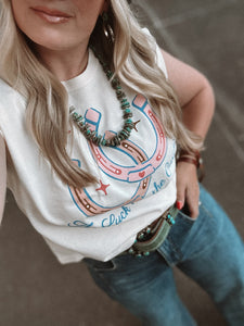 Person wearing a white tank top with colorful designs and blue jeans, standing on a street.