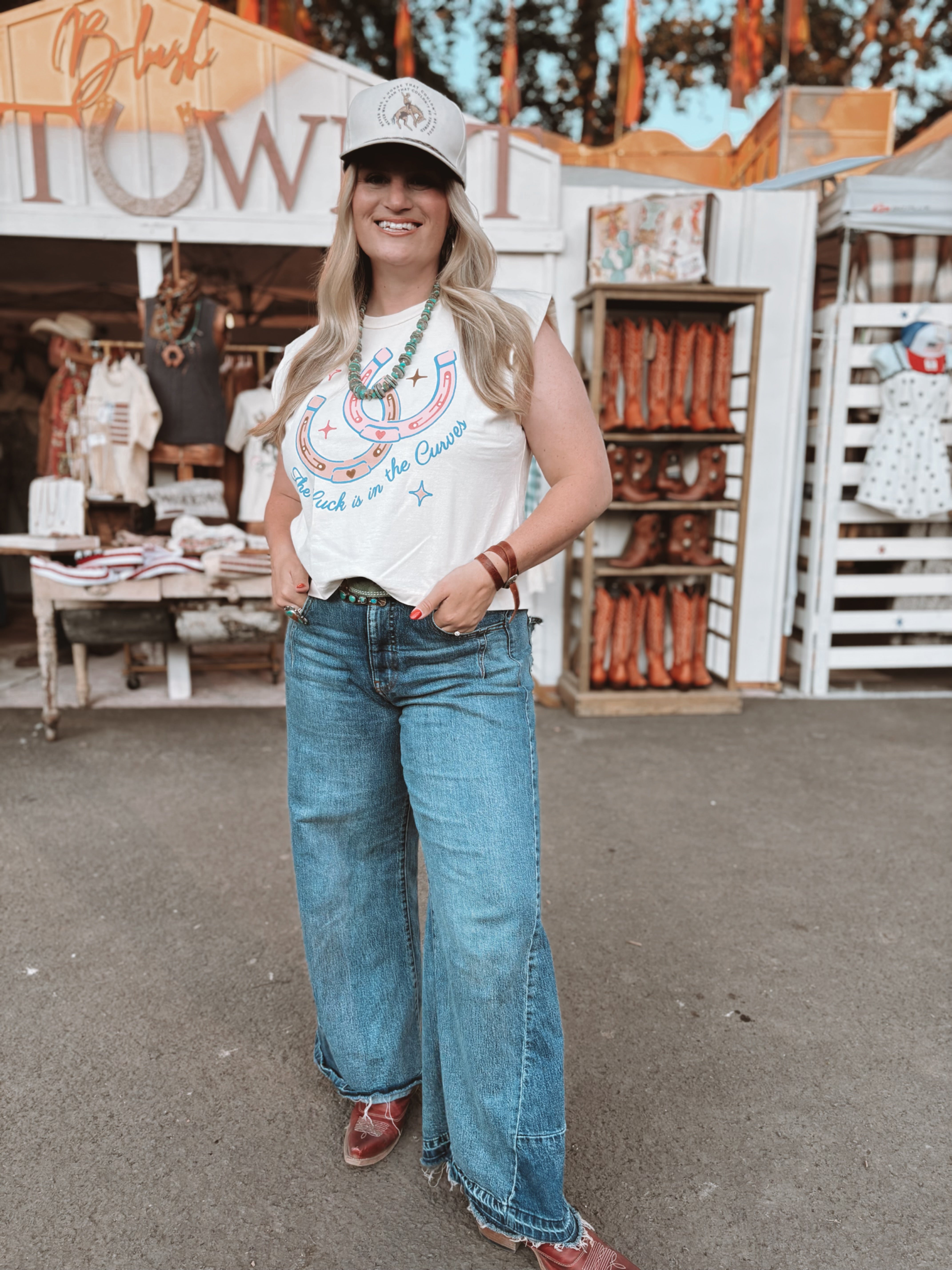 Woman in a white graphic tank top and blue jeans standing in front of a store with cowboy boots.