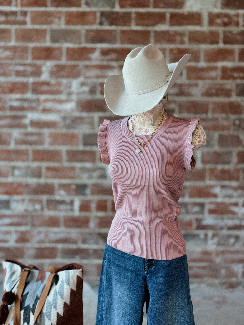 Mannequin wearing a pink sleeveless top and blue jeans with a cowboy hat against a brick wall.