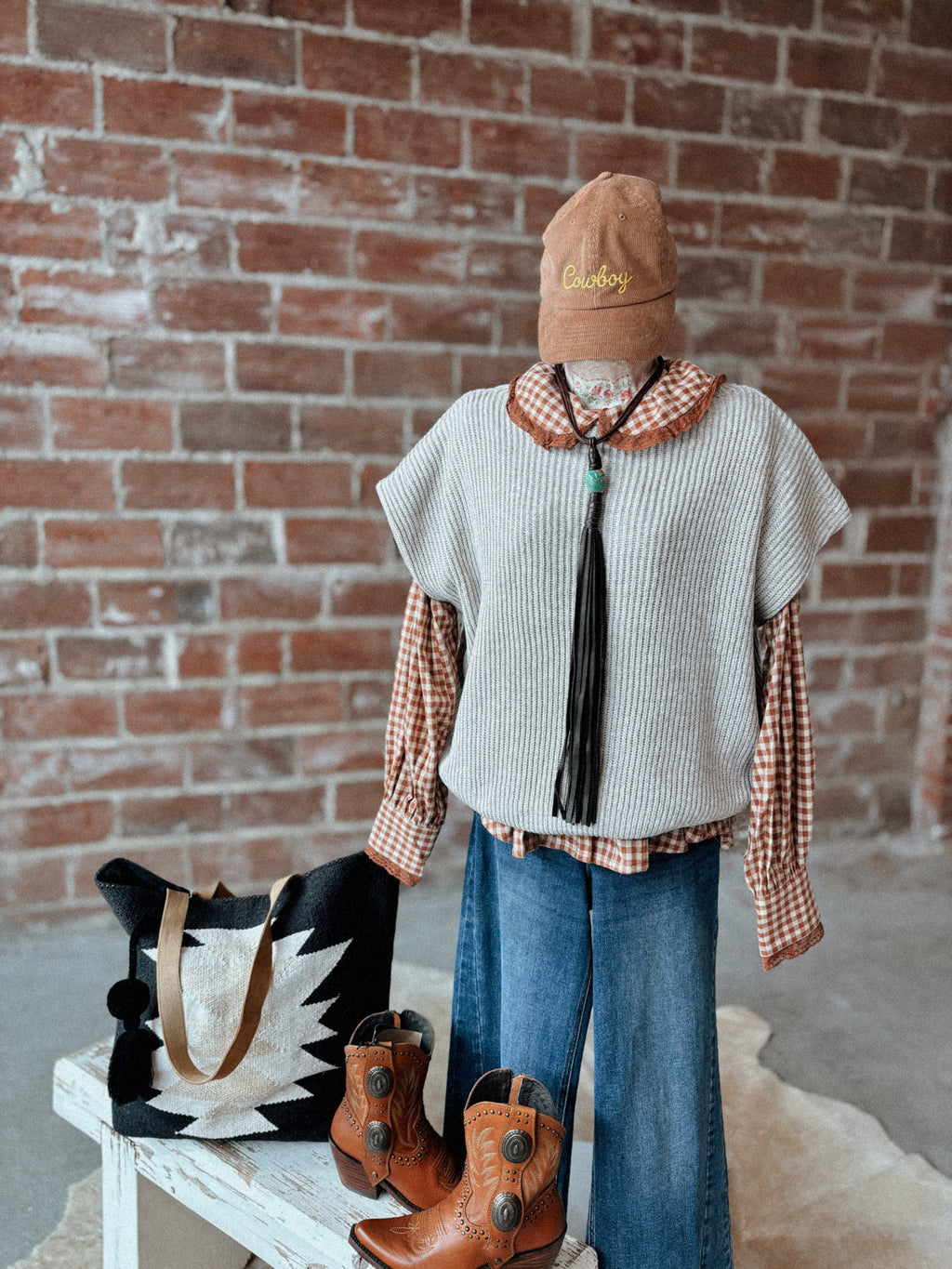 Mannequin wearing a stylish outfit with a hat, shirt, vest, jeans, and boots against a brick wall.