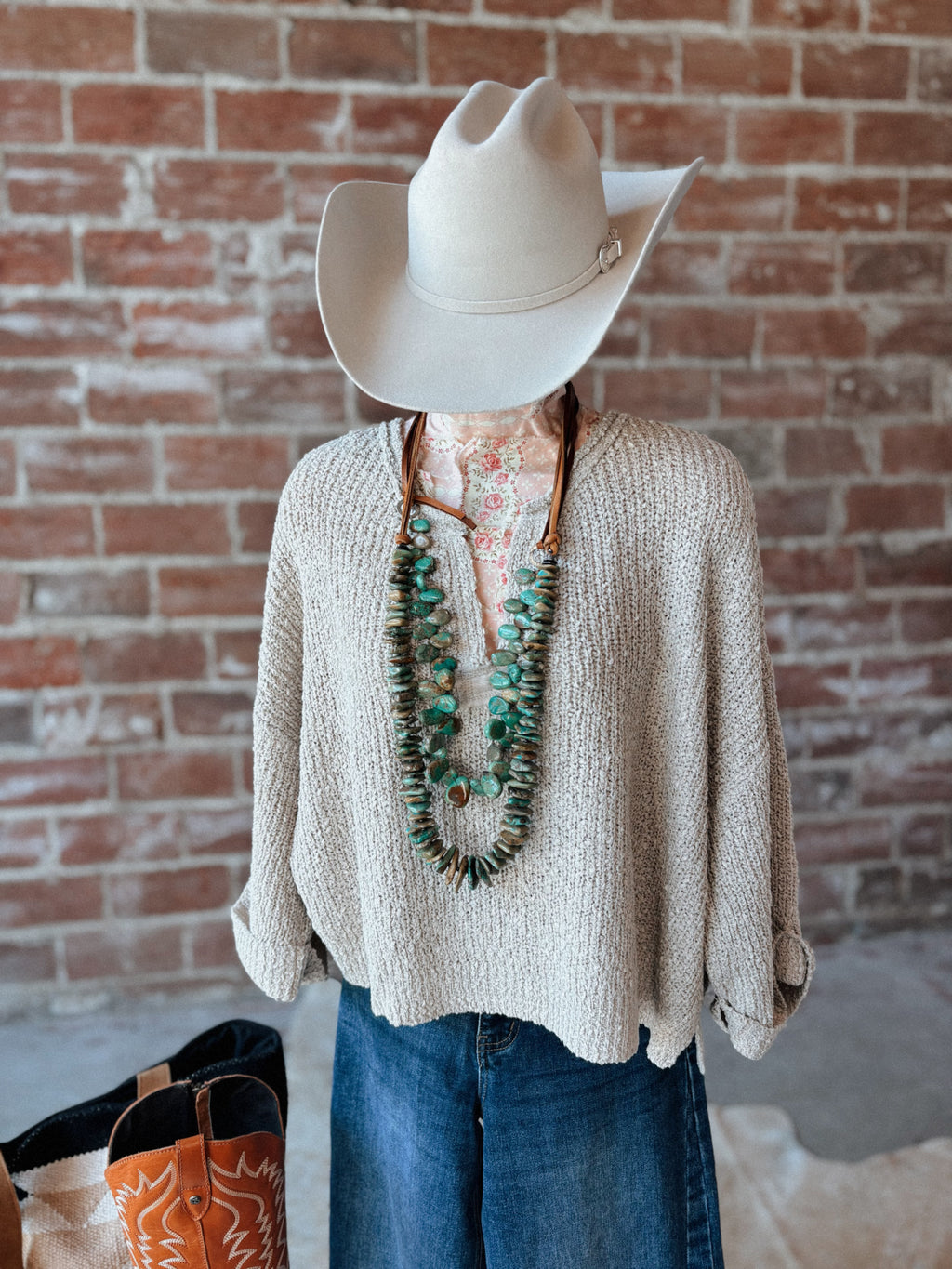 Mannequin wearing a beige sweater, blue jeans, and a white cowboy hat against a brick wall.