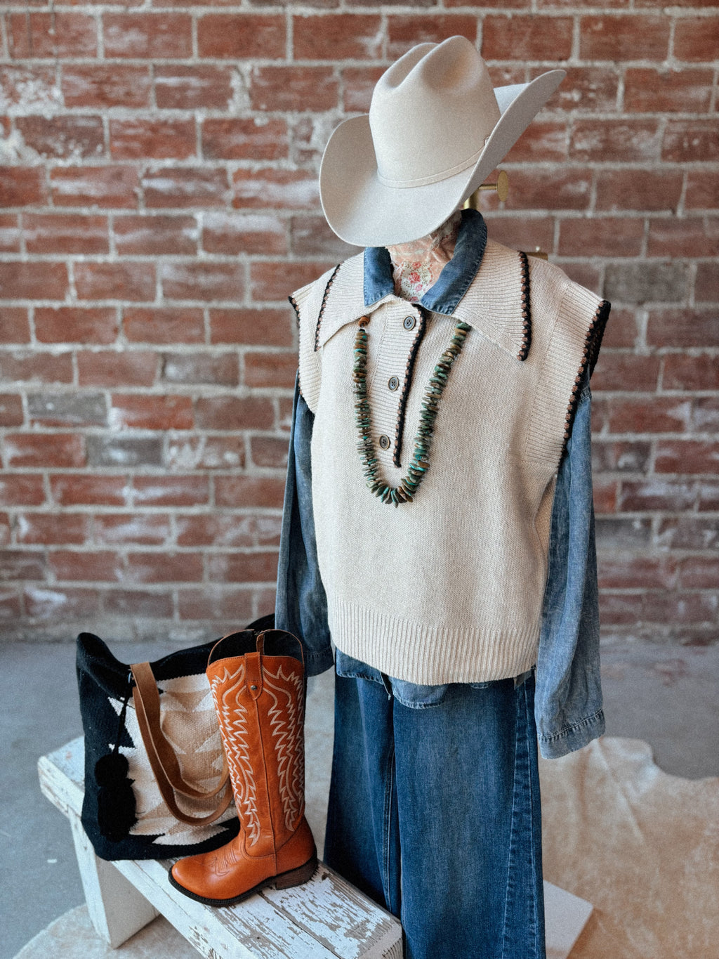 Mannequin wearing a beige vest, blue shirt, and cowboy hat with orange boots against a brick wall.