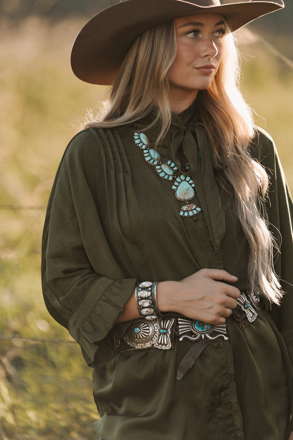 Woman in a green outfit with a cowboy hat and jewelry in a natural setting