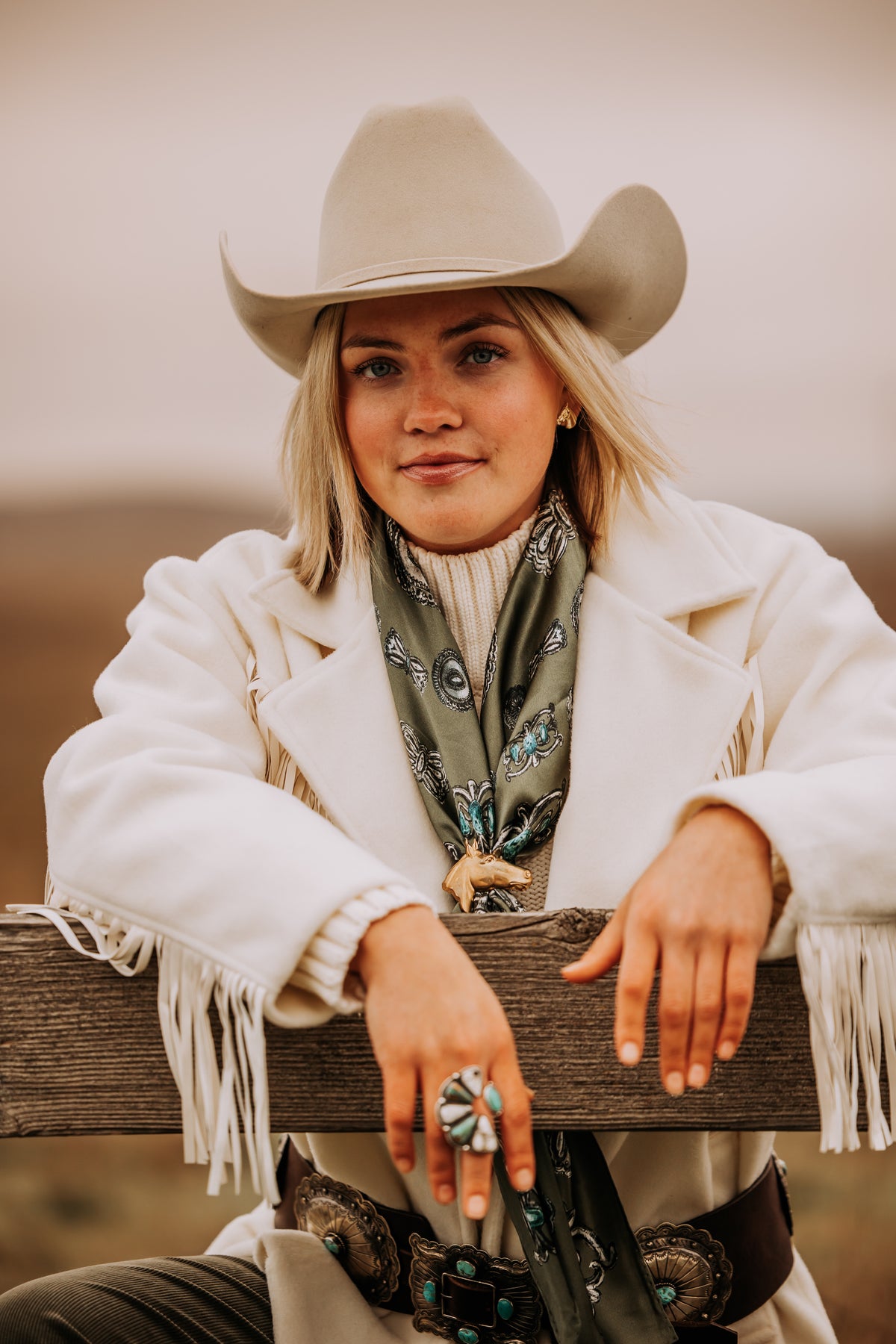 A woman in a cowboy hat and cream fringe jacket wearing an olive wild rag