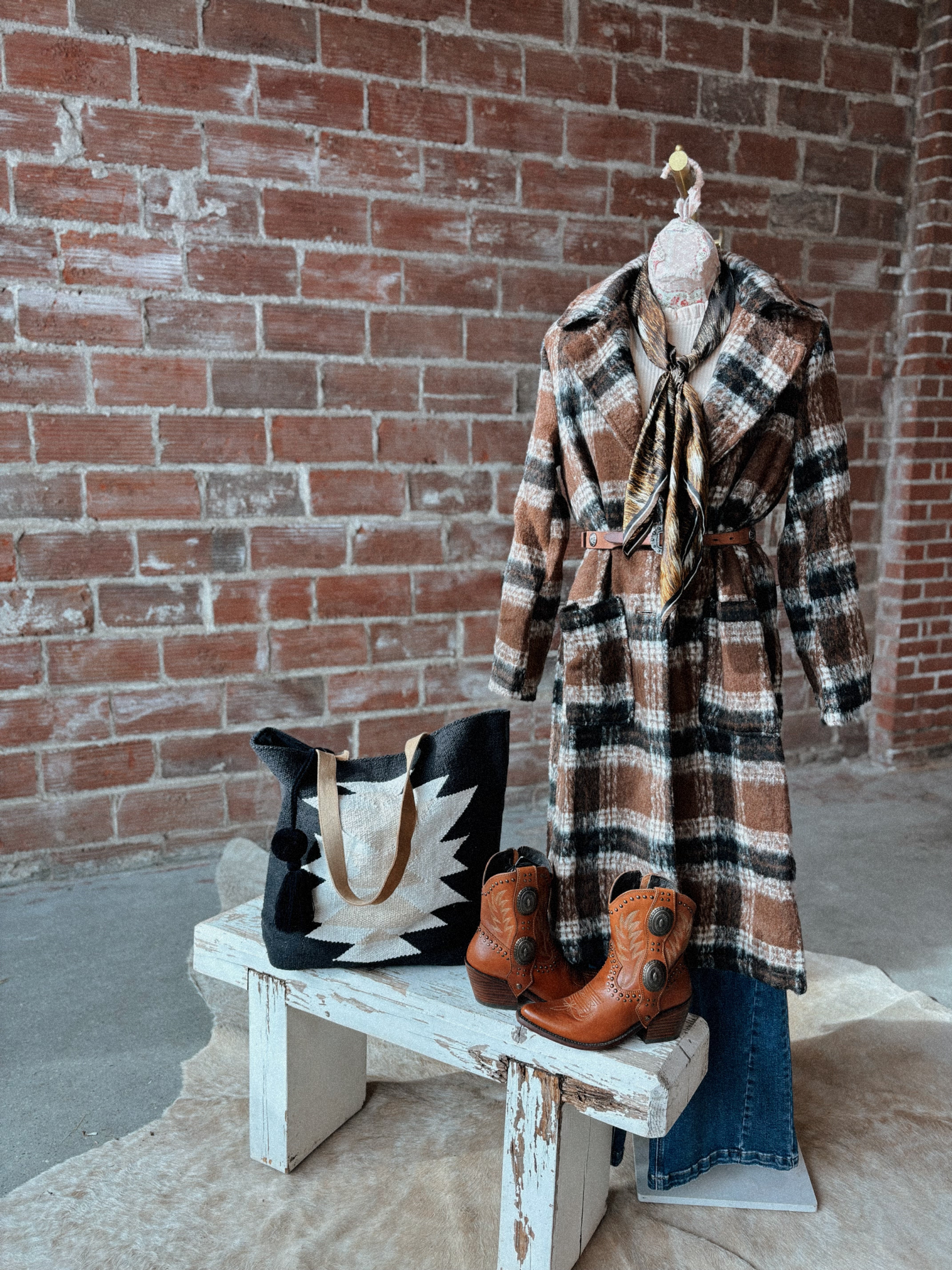 Plaid coat, boots, and handbag on a wooden bench against a brick wall.