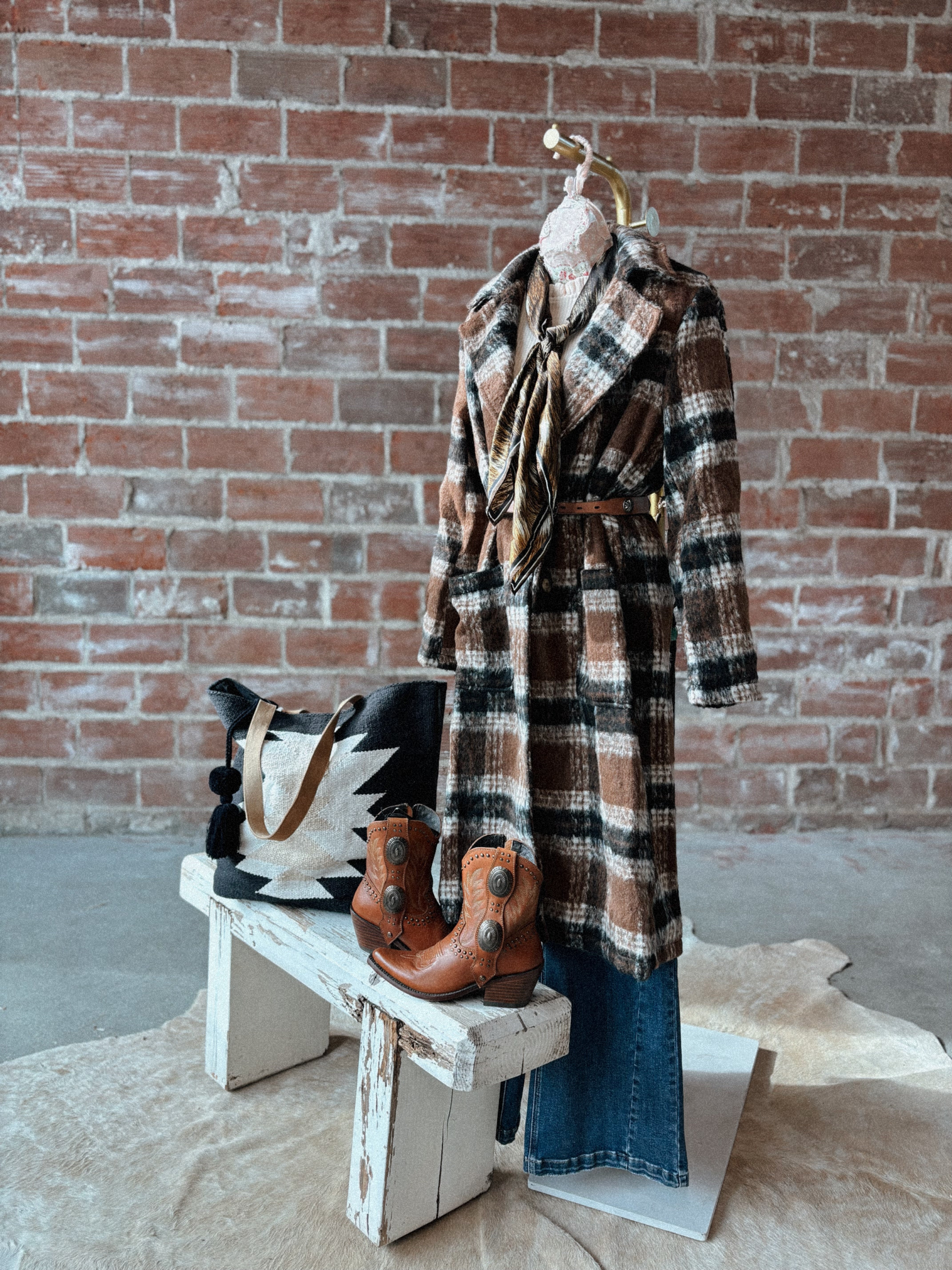 Plaid coat on a mannequin with boots and a bag on a wooden bench against a brick wall.