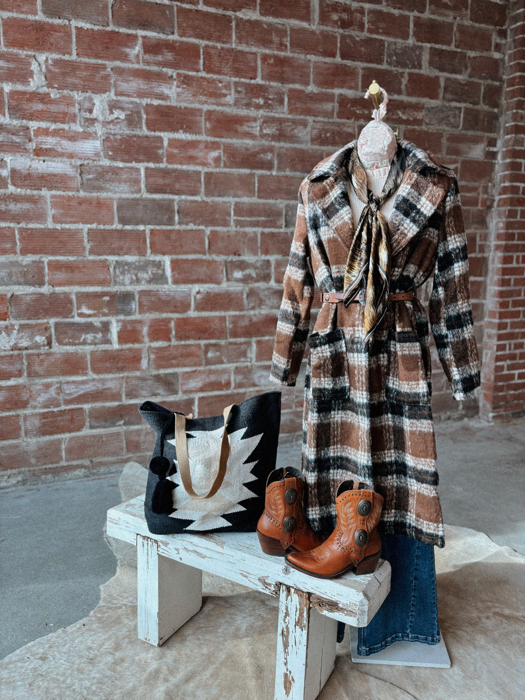 Plaid coat, boots, and handbag on a wooden bench against a brick wall.