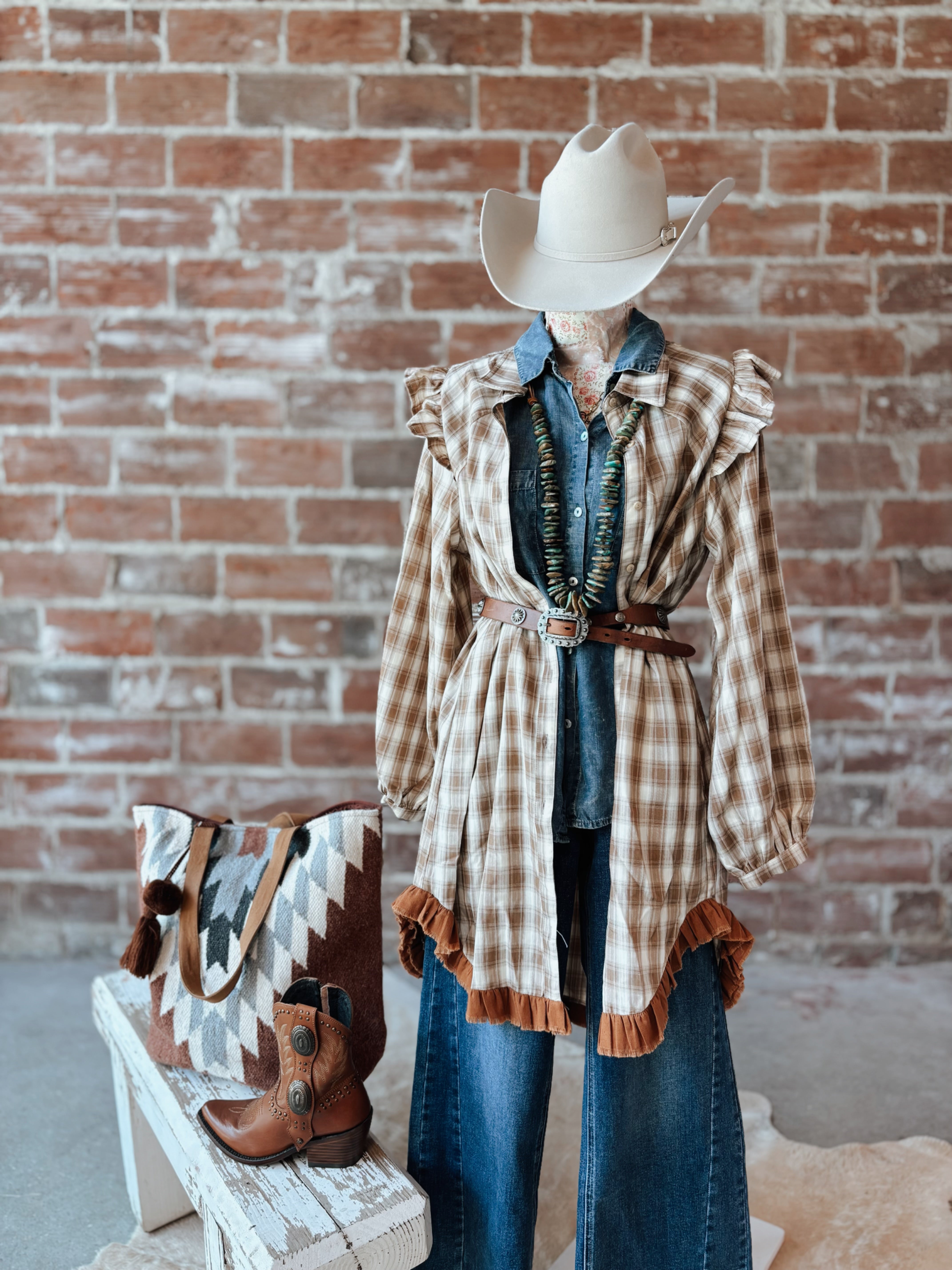 Mannequin wearing a plaid shirt, denim jacket, and cowboy hat with a patterned bag against a brick wall.