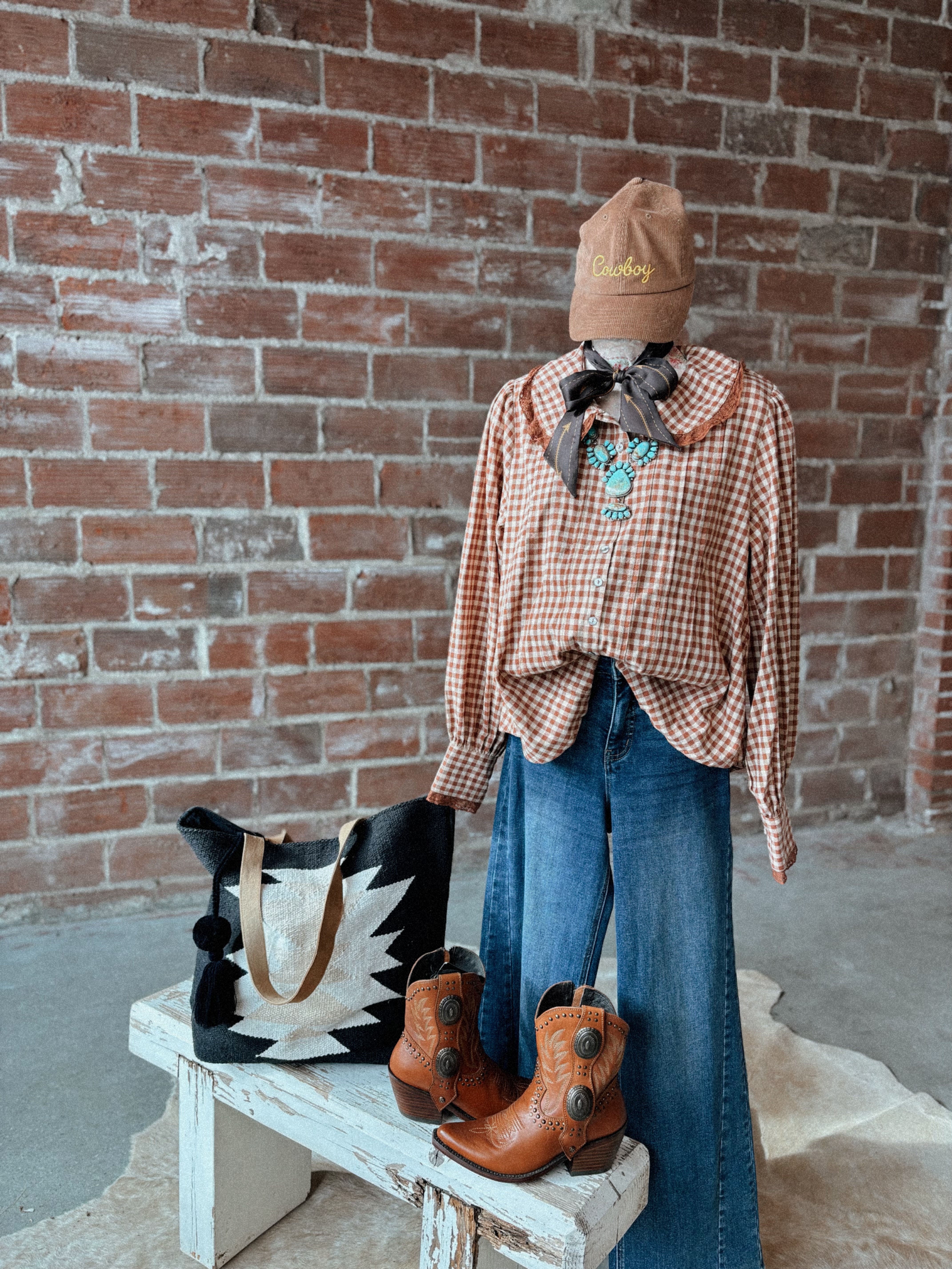 Mannequin wearing a plaid shirt, blue jeans, and brown boots against a brick wall.