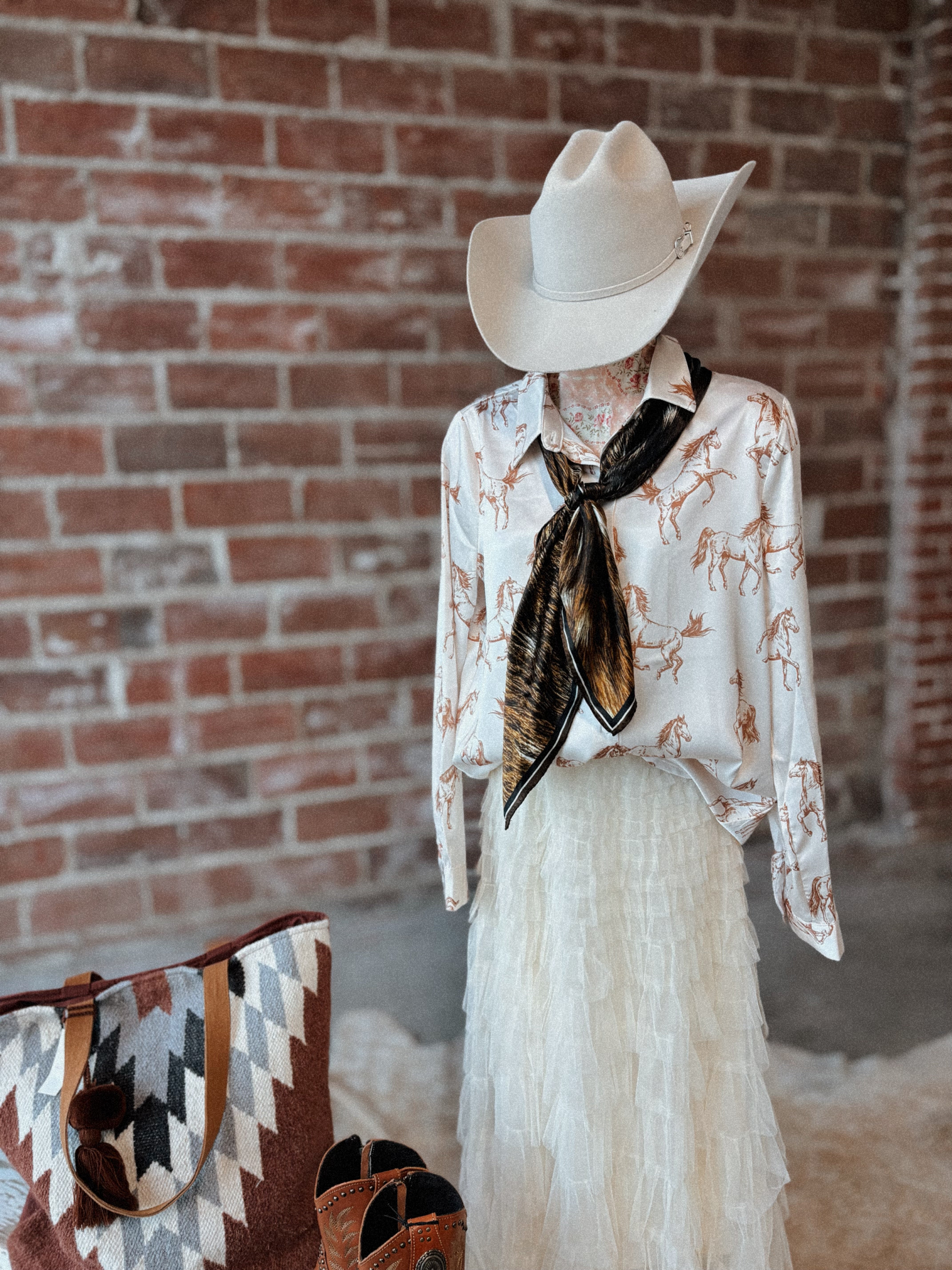 Mannequin wearing a cowboy hat, patterned shirt, and white skirt against a brick wall.