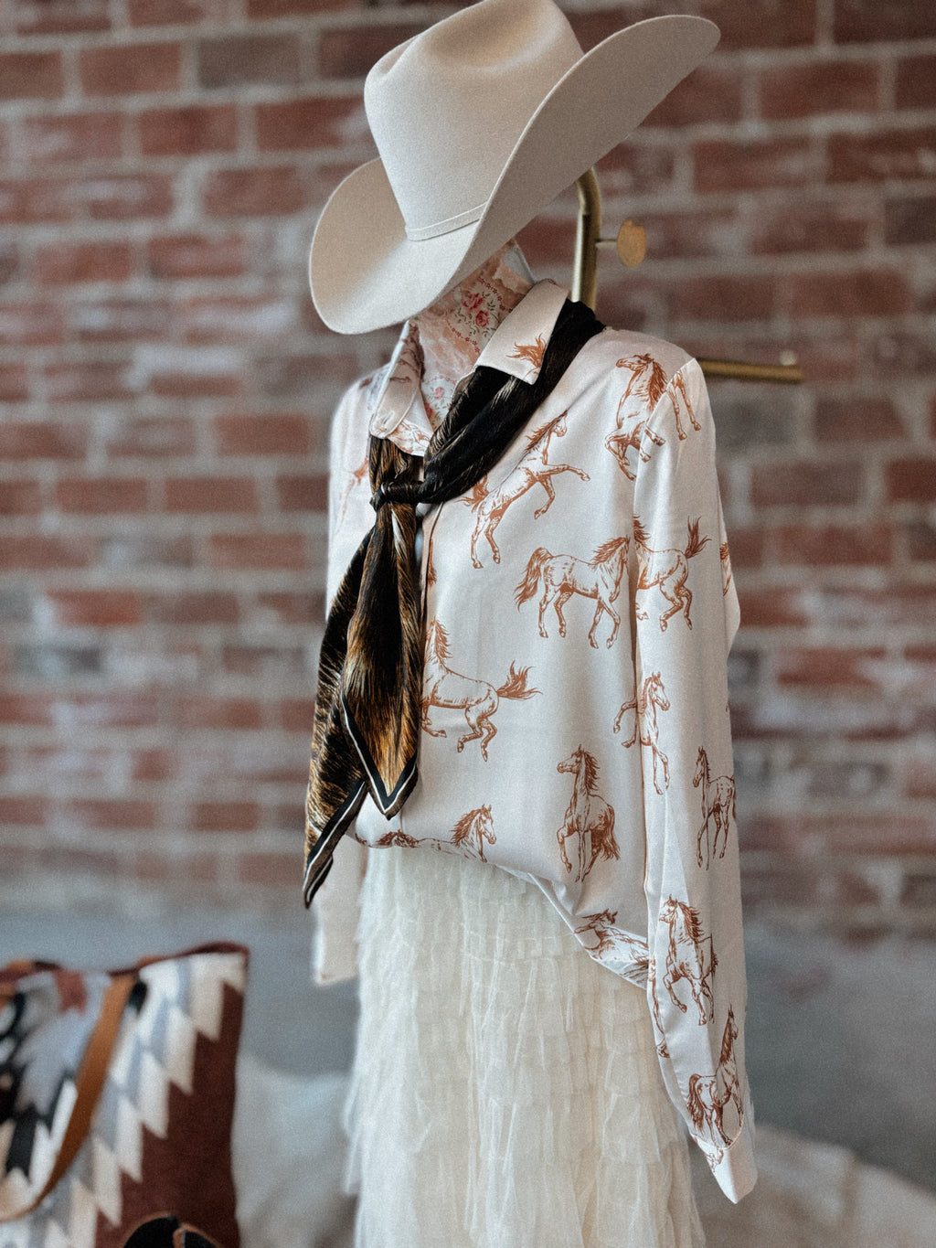 Mannequin wearing a white blouse with horse prints, a white cowboy hat, and a patterned scarf against a brick wall.