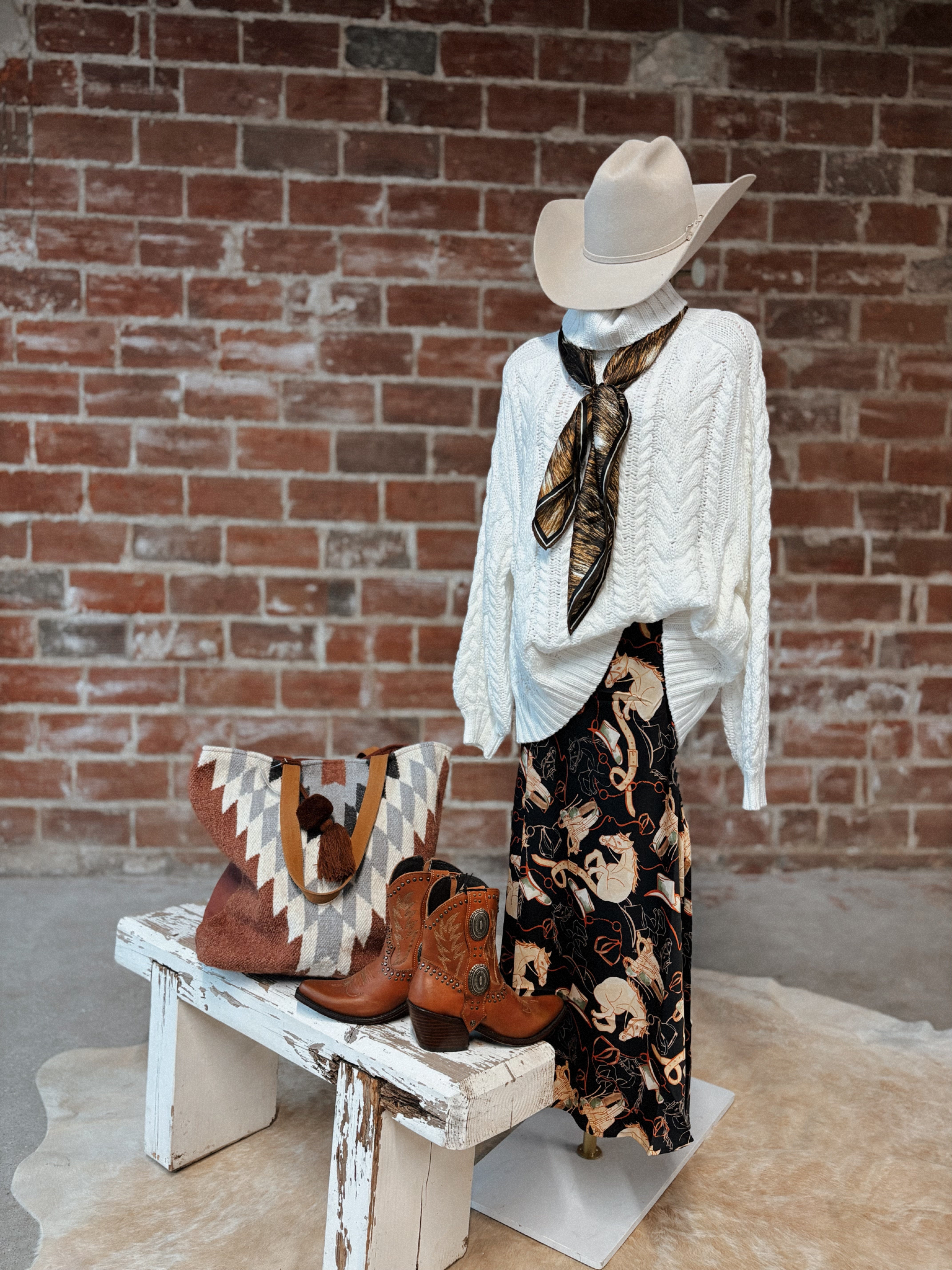Mannequin wearing a white sweater, patterned skirt, and cowboy hat with boots and handbag on a wooden bench against a brick wall.
