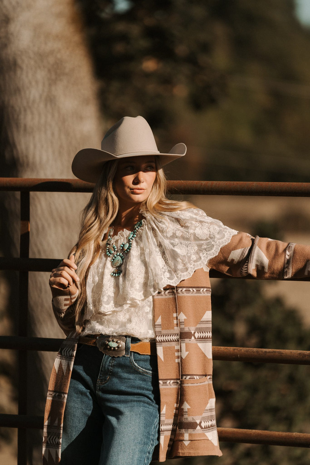 Person wearing a cowboy hat and patterned coat standing against a wooden fence with trees in the background.