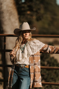 Person wearing a cowboy hat and patterned coat standing against a wooden fence with trees in the background.
