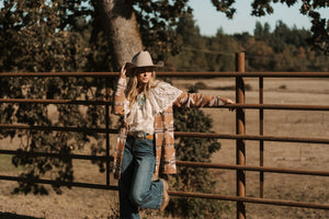 Person wearing a cowboy hat and plaid shirt leaning against a wooden fence in a rural setting.