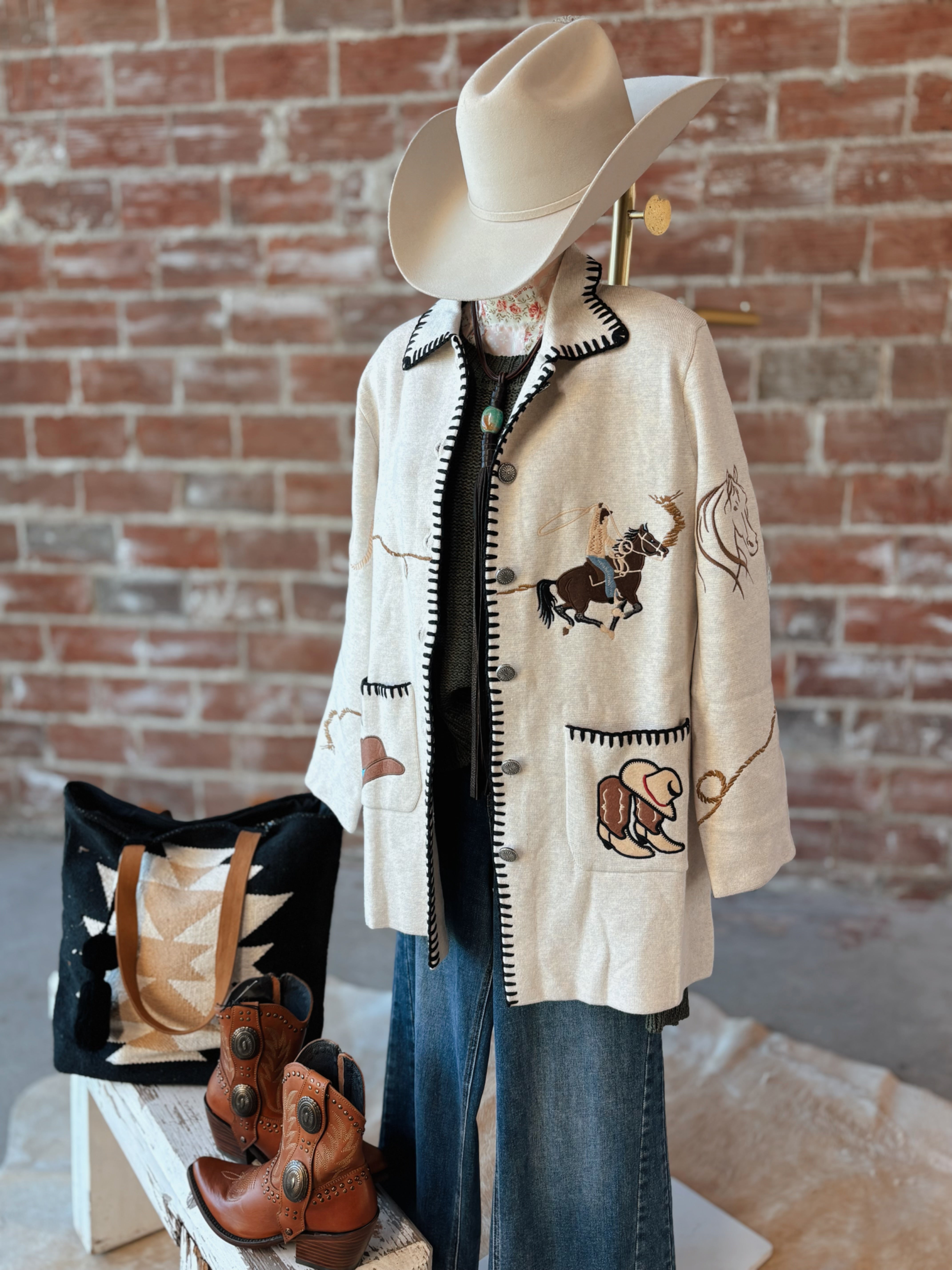 Mannequin wearing a western-style coat with bull design, cowboy hat, and boots against a brick wall.