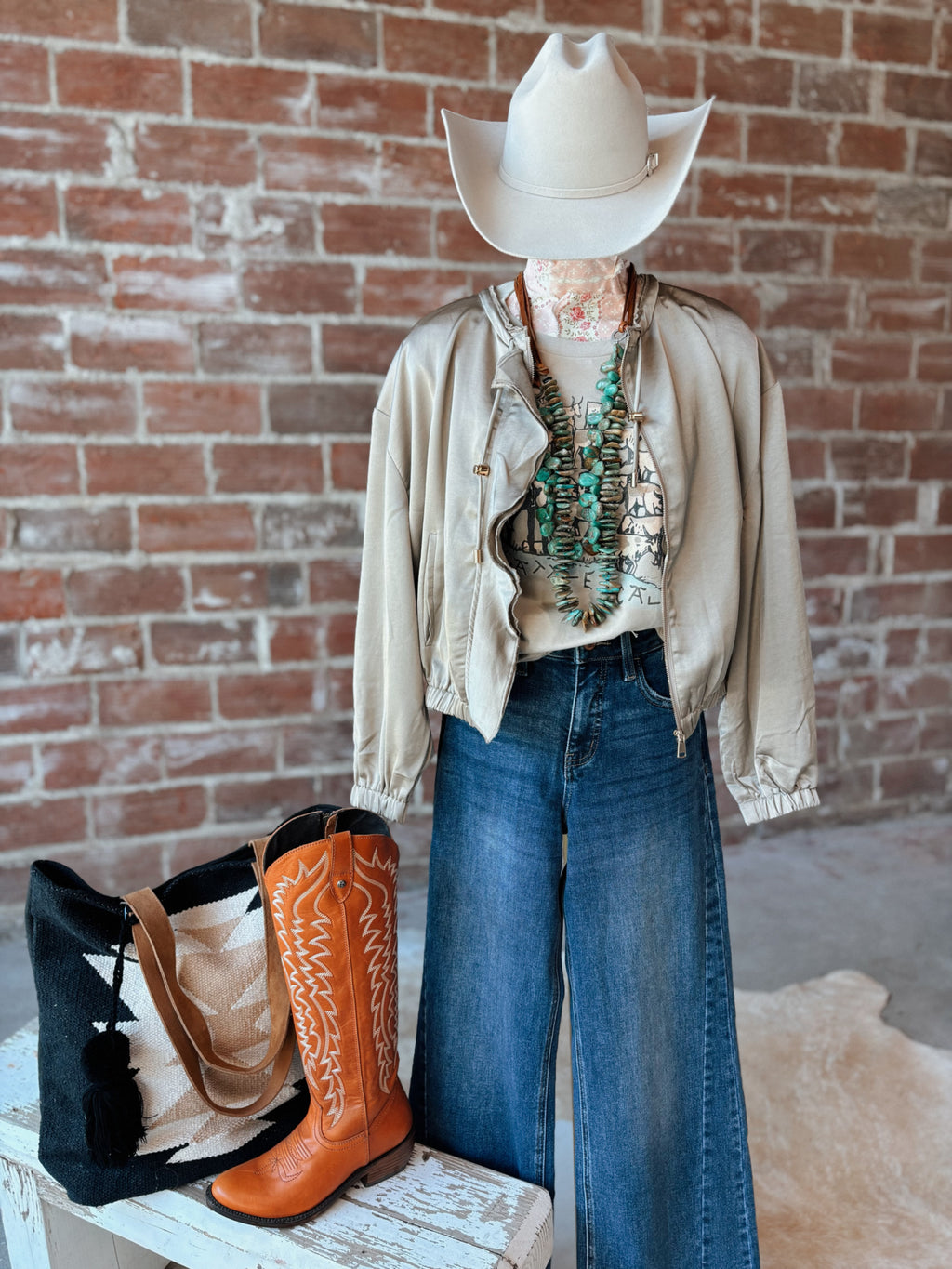 Person wearing a beige jacket, blue jeans, and orange cowboy boots with a white cowboy hat against a brick wall.